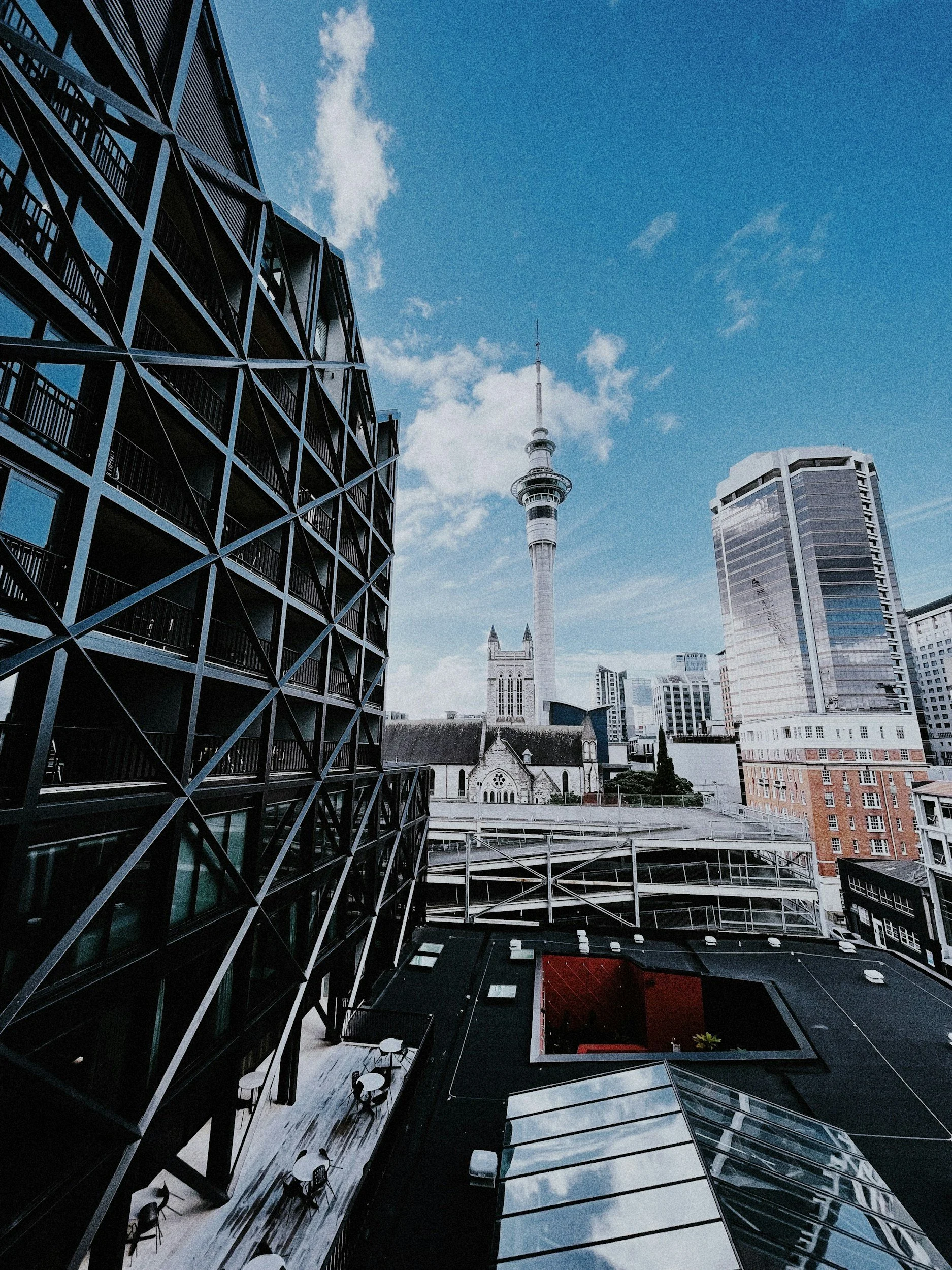 A cityscape showing modern and historic buildings, including the Sky Tower, under a partly cloudy sky.