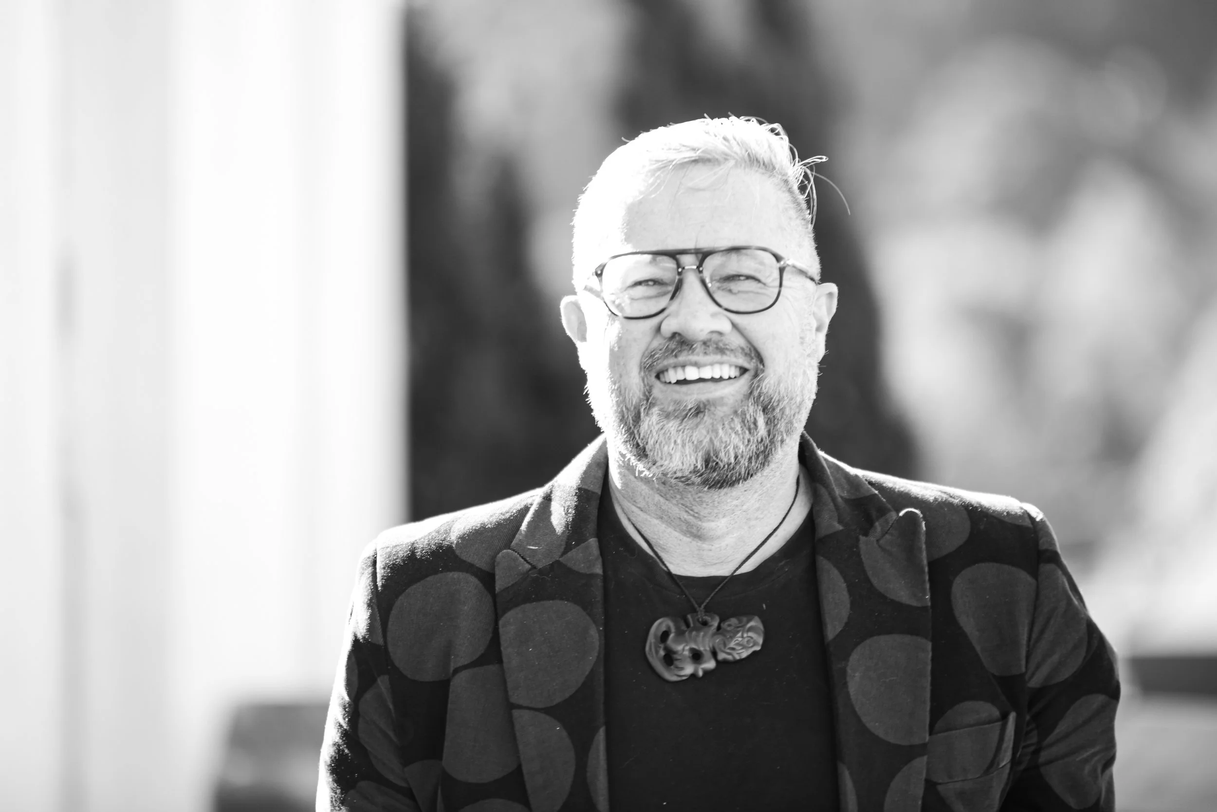 A smiling man with glasses, facial hair, and short light hair, wearing a patterned jacket over a dark shirt, standing outdoors.