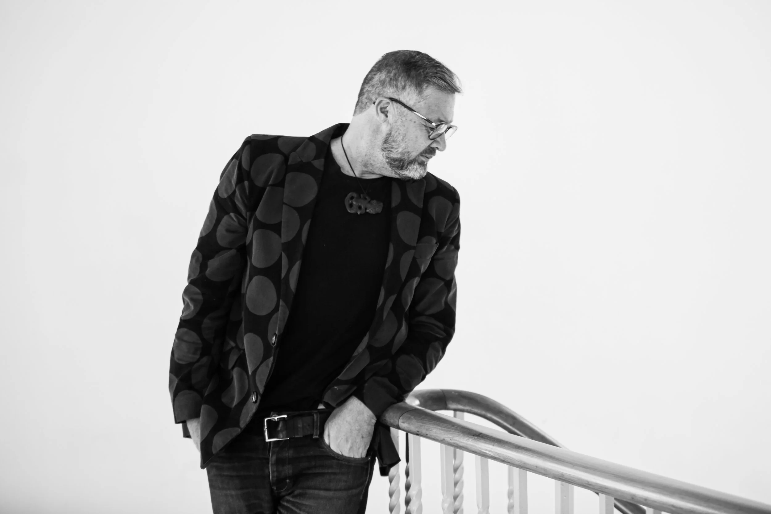 A man with glasses, a beard, and short hair is leaning on a railing, looking down. He is wearing a polka dot blazer over a black shirt, and a necklace with a skull pendant. The image is in black and white and has a plain white background.