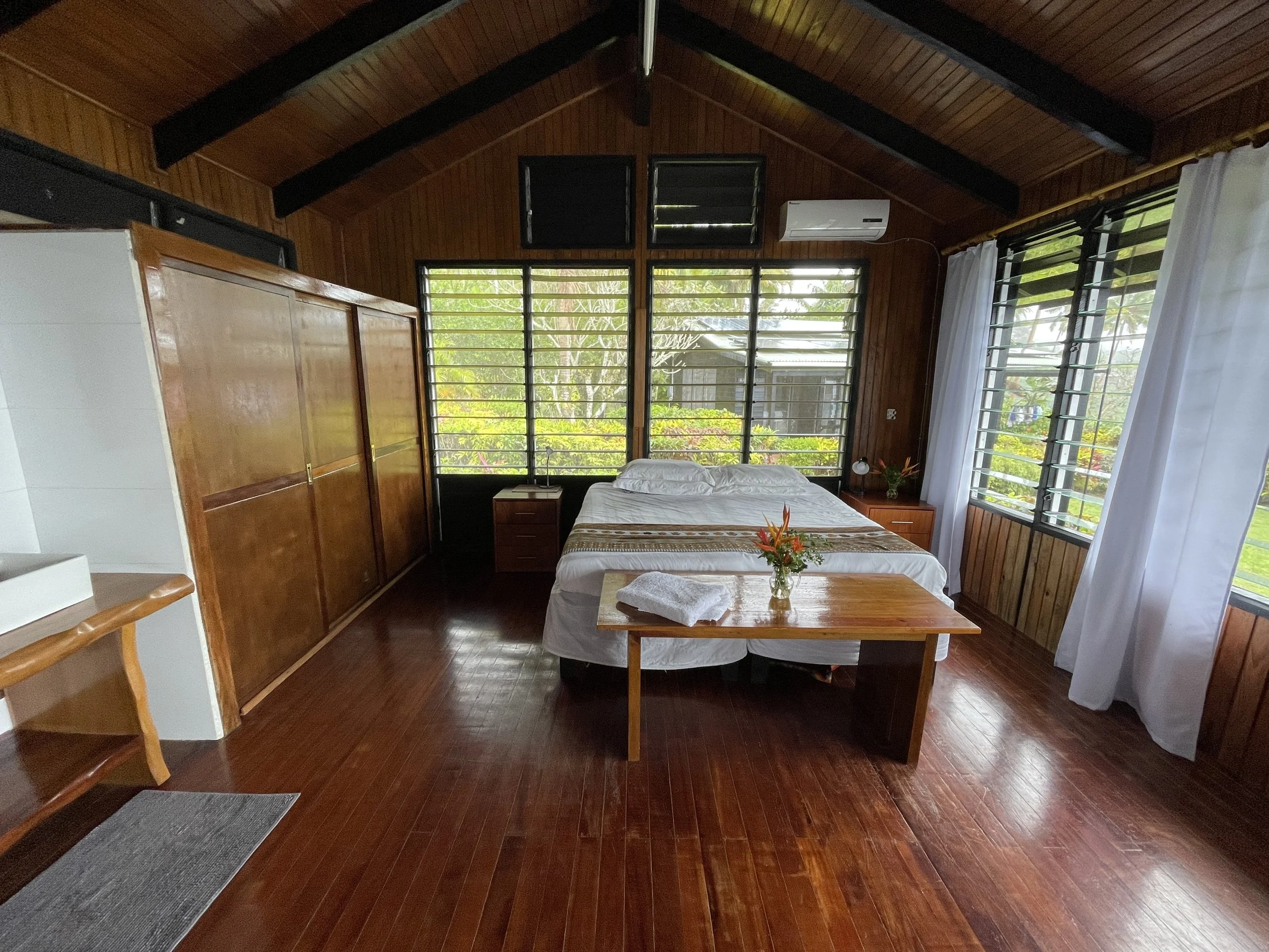 A cozy bedroom with wooden walls and ceiling, featuring a bed with white linens, a wooden side table with a small lamp, a flower vase, and windows with black frames and white curtains, overlooking lush greenery outside.