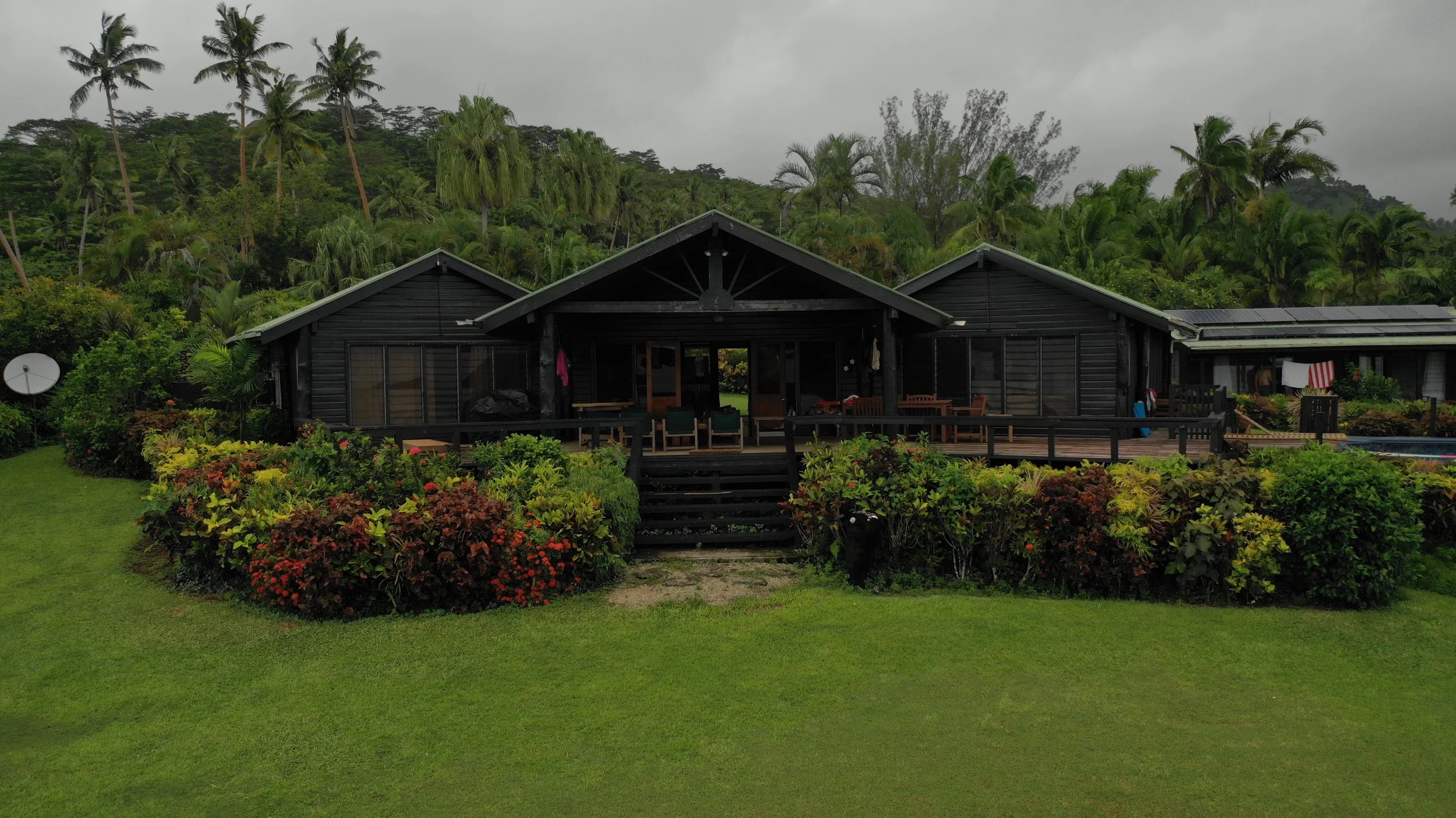 A black wooden house with a porch and landscaped garden, surrounded by lush green trees and hills, on a cloudy day.