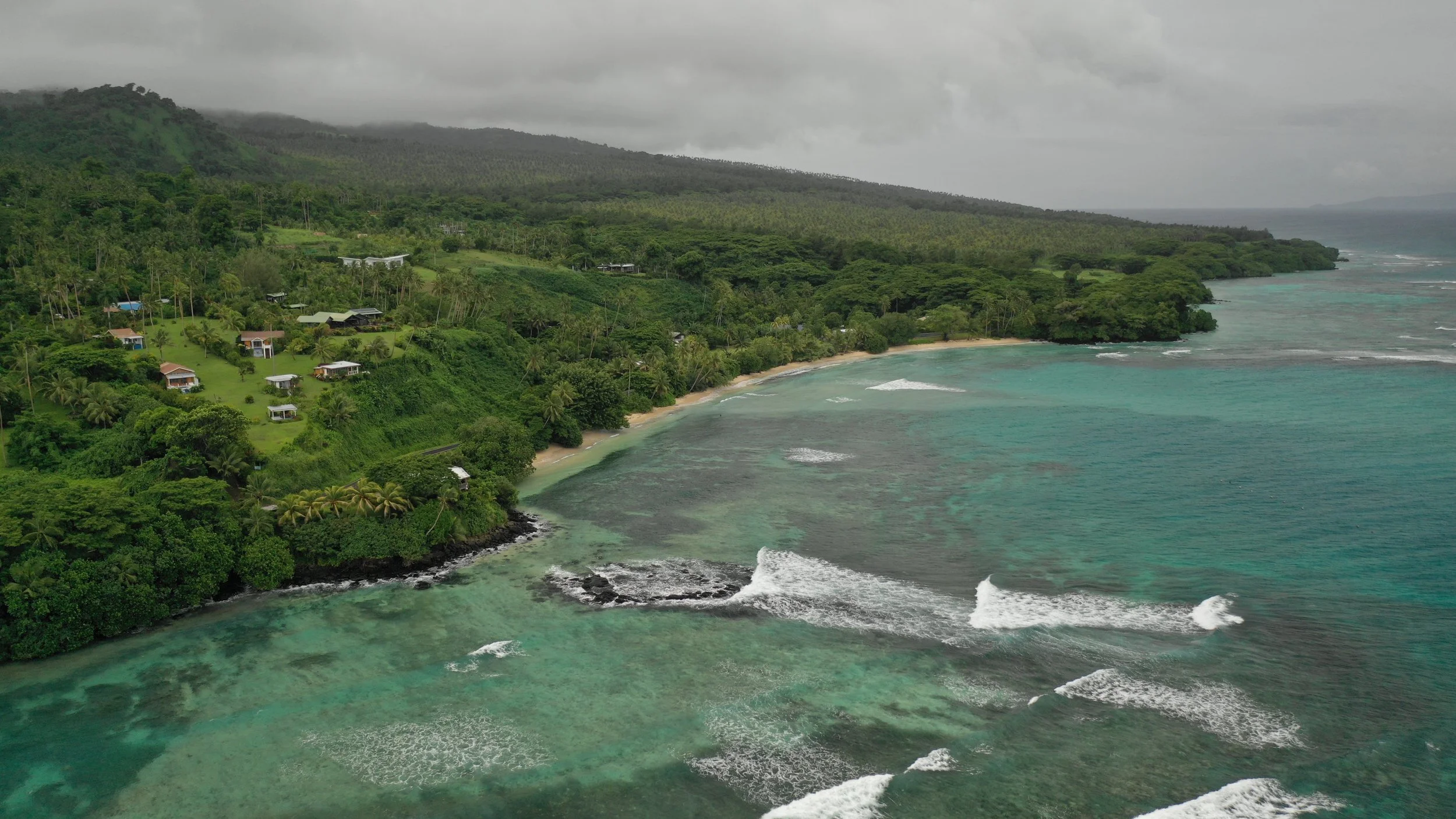 Ucuilagi, Taveuni Island Fiji - Aerial view of a lush green coastline with tropical trees and houses, turquoise ocean waves crashing onto a small sandy beach, and mountains covered in clouds in the background.