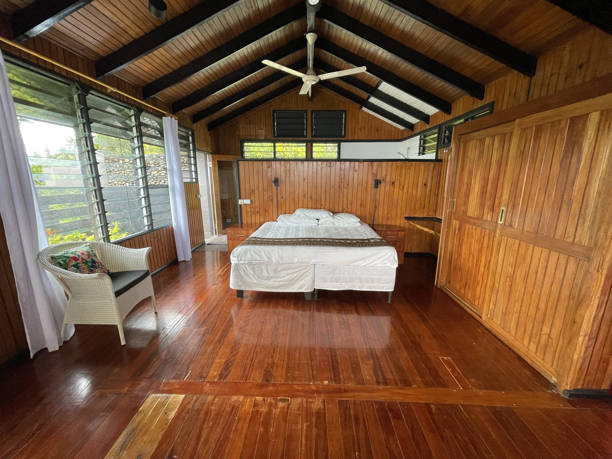 Ucuilagi, Taveuni Island Fiji - Master bedroom with wooden walls and ceiling, large louvered windows, a bed with white bedding, a wicker chair with a floral cushion, and a ceiling fan.