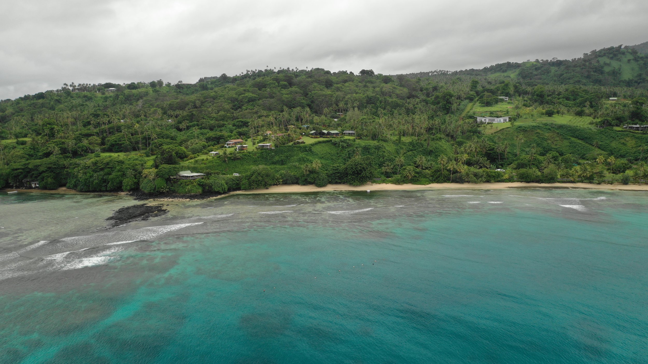 A lush green hillside with houses and trees overlooking a sandy beach and turquoise ocean under cloudy skies.