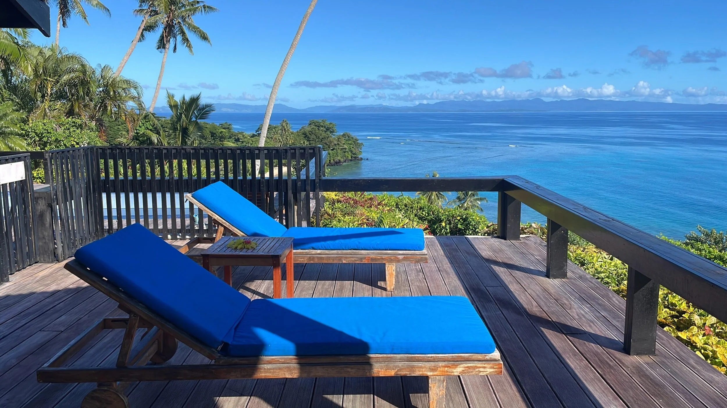 Ucuilagi, Taveuni Island Fiji - A wooden deck with two blue lounge chairs and a small table, overlooking a tropical landscape with lush green trees, palm trees, and a view of the ocean and distant mountains under a blue sky with clouds.