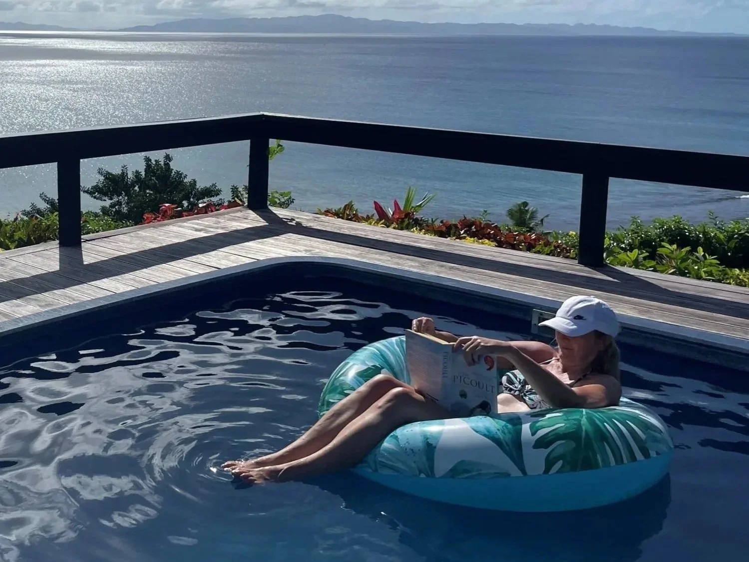 Ucuilagi, Taveuni Island Fiji - A woman relaxing in a pool on an inflatable ring, reading a book, with ocean view and deck railing in the background.