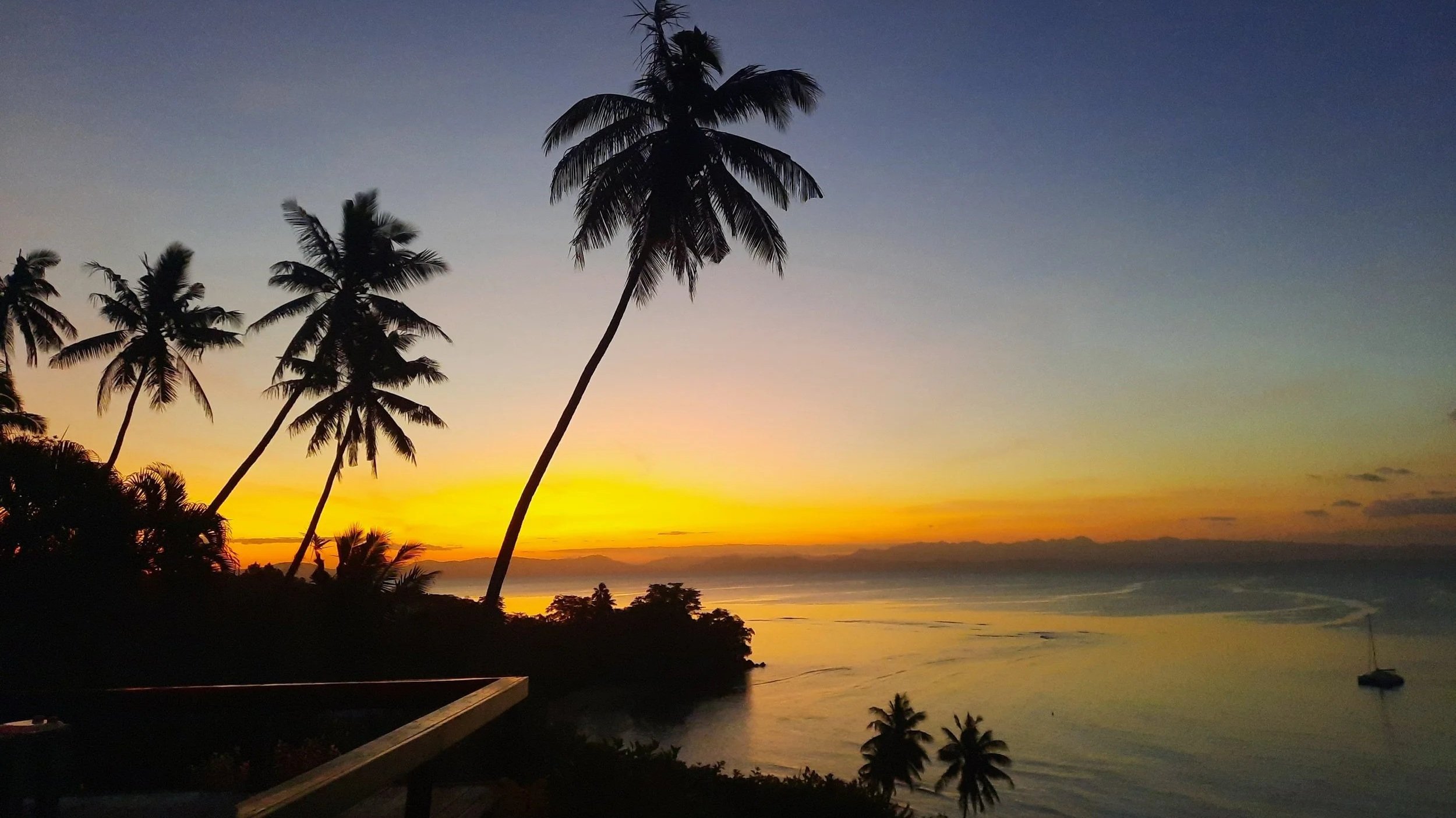 Ucuilagi, Taveuni Island Fiji - View from the mainhouse - Silhouettes of palm trees against a colorful sunset sky with shades of yellow, orange, and purple over a coastal landscape.