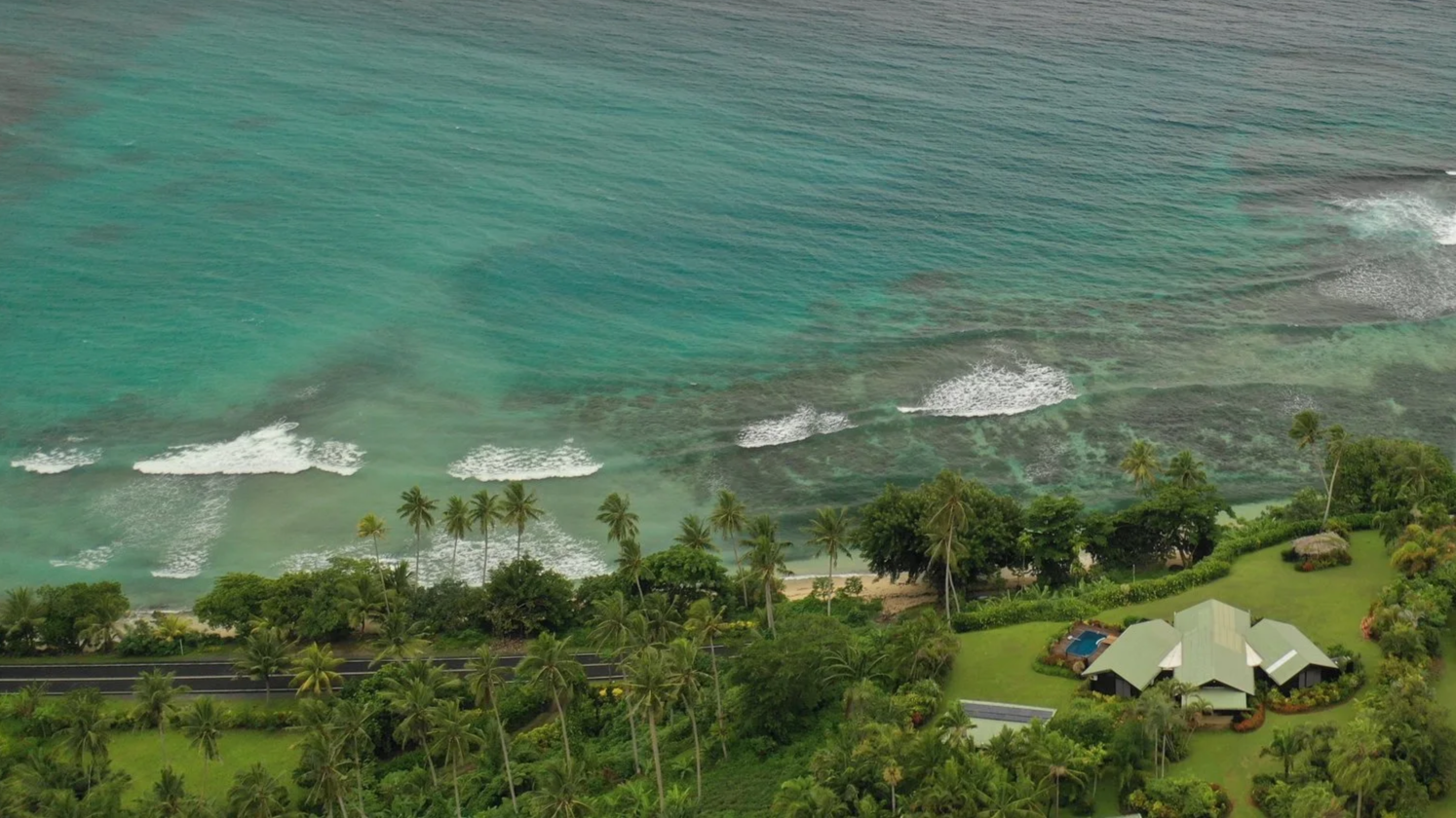 View of a tropical coastline with clear turquoise water, white waves, lush green trees, and several houses along the shore under a cloudy sky.