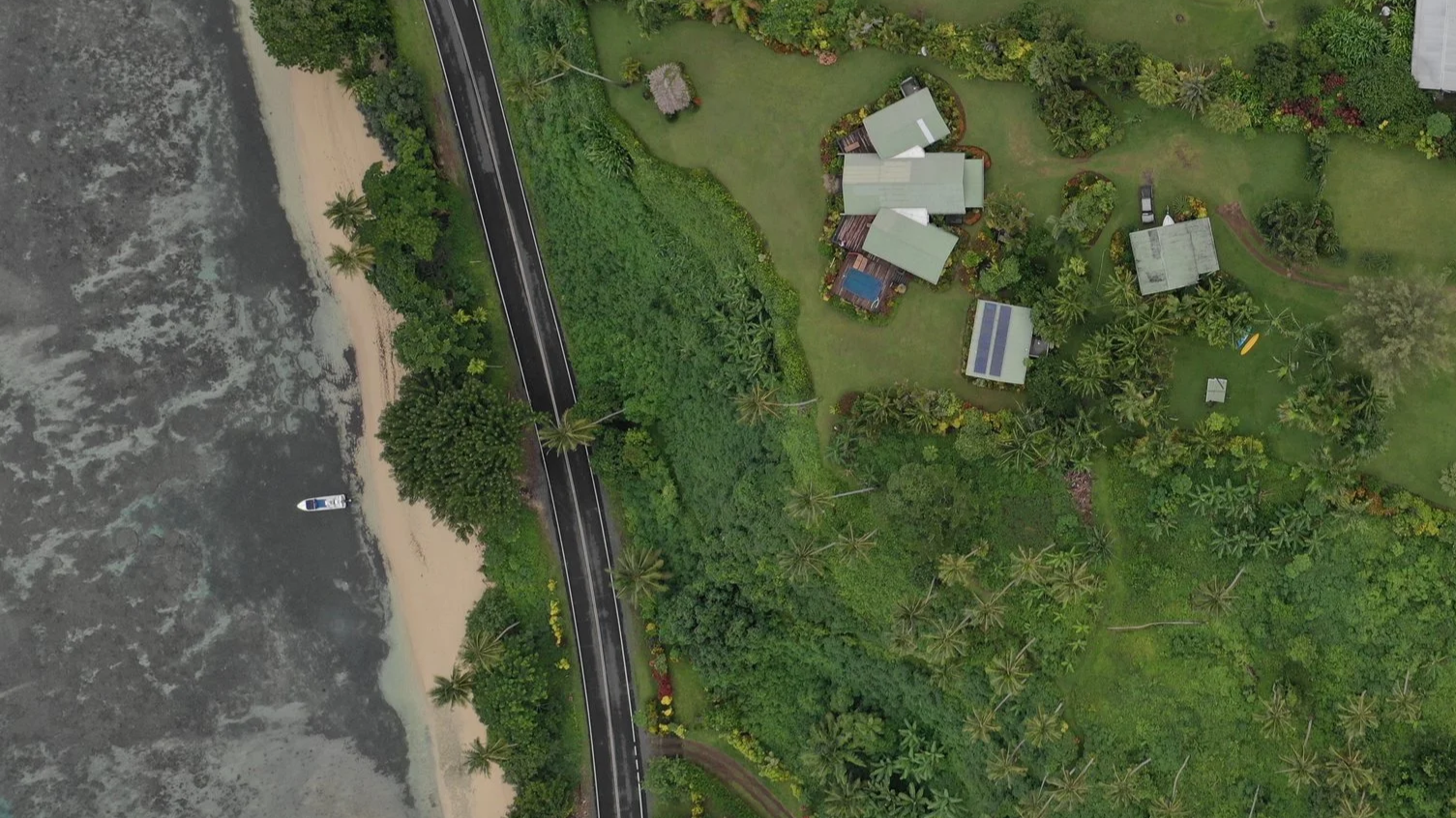 Aerial view of a coastal property with several houses, lush greenery, palm trees, and a coastline with a beach and ocean waves.