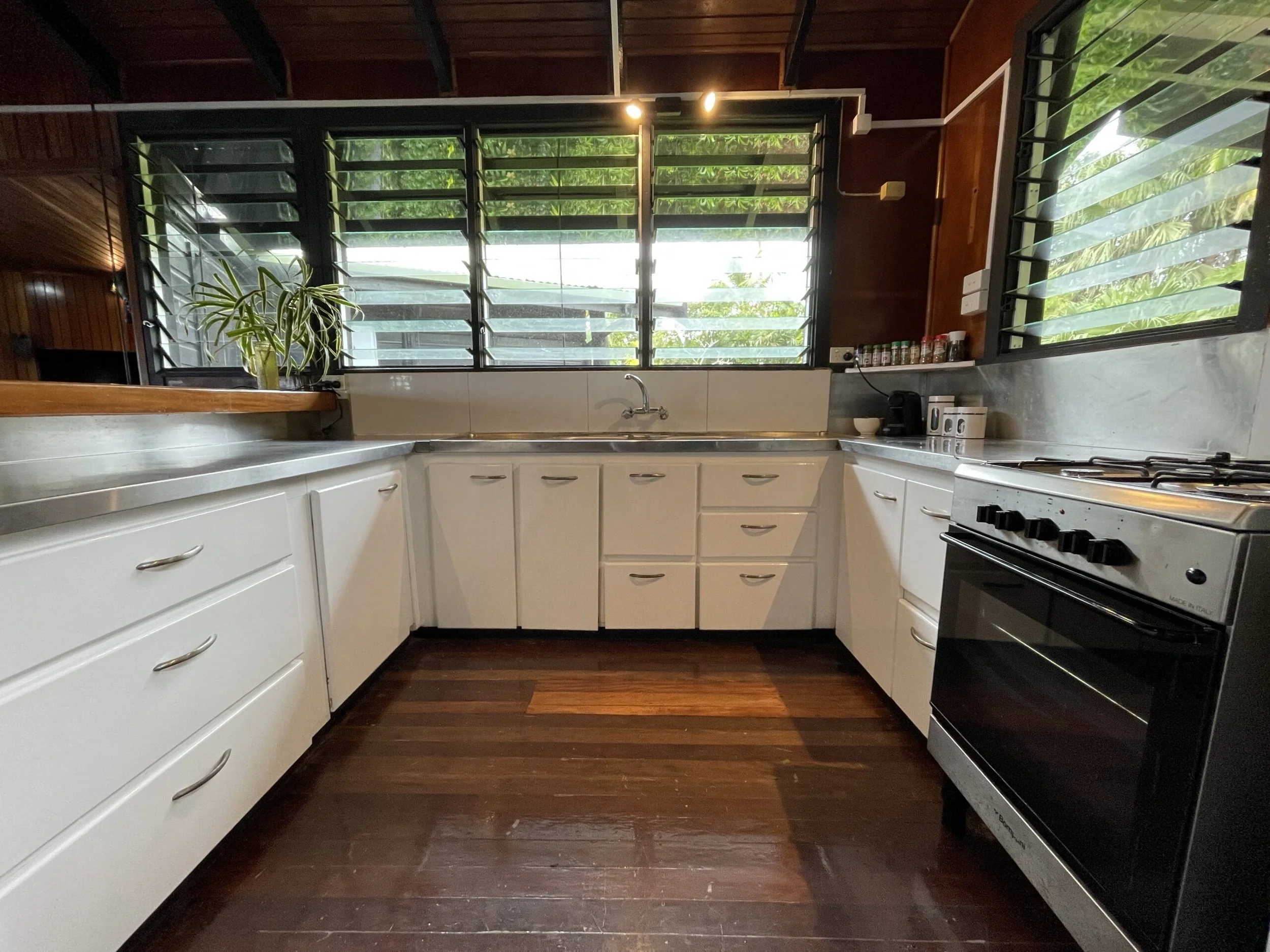 Ucuilagi, Taveuni Island Fiji - Kitchen with white cabinets, dark wood flooring, large window with adjustable black shutters above the sink, a plant on the left side of the window, spices on the right side, and a stove on the right.