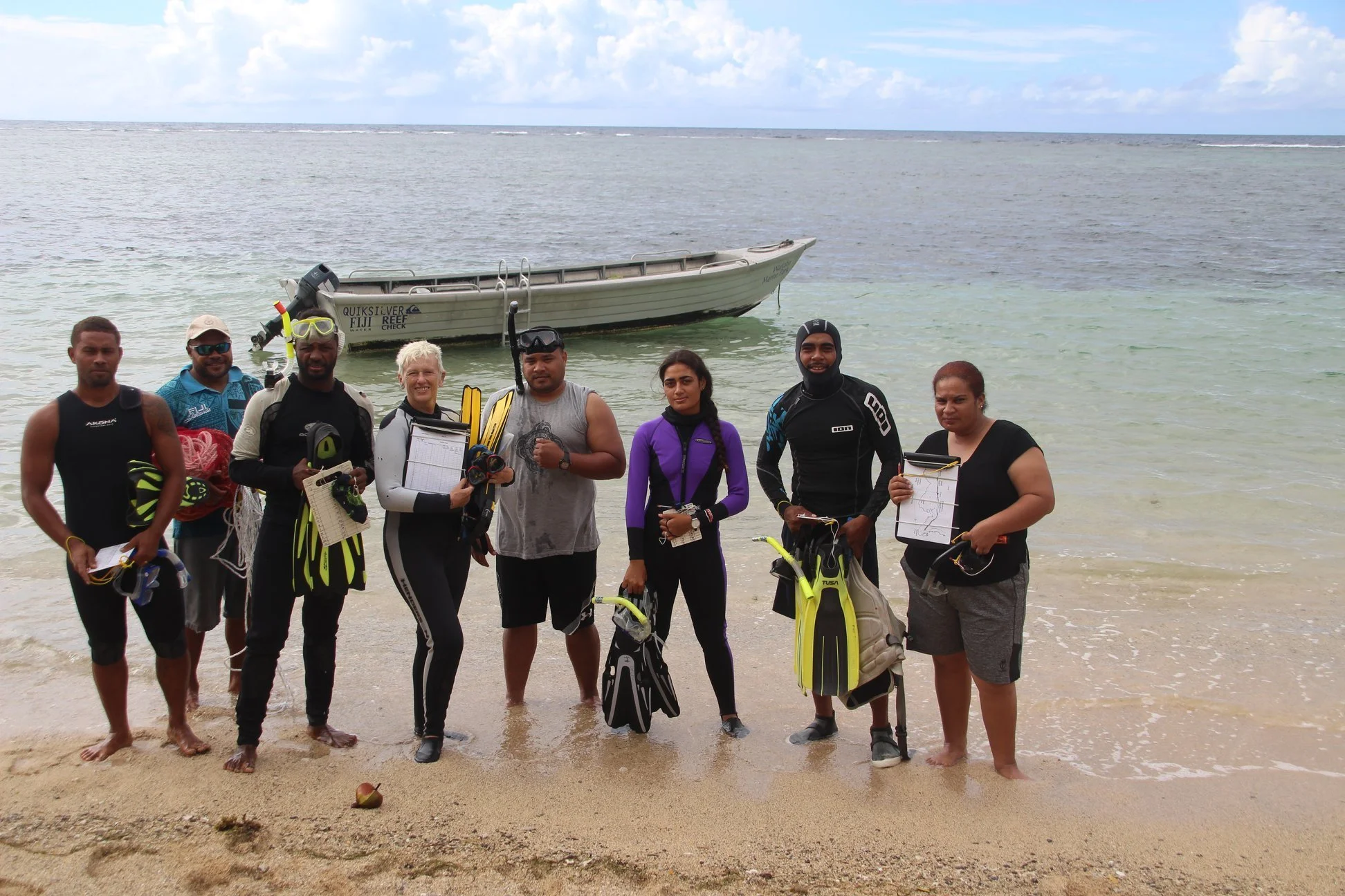 Group of eight people standing on a beach with a boat in the background, some holding diving or snorkeling gear, near the water during daytime.