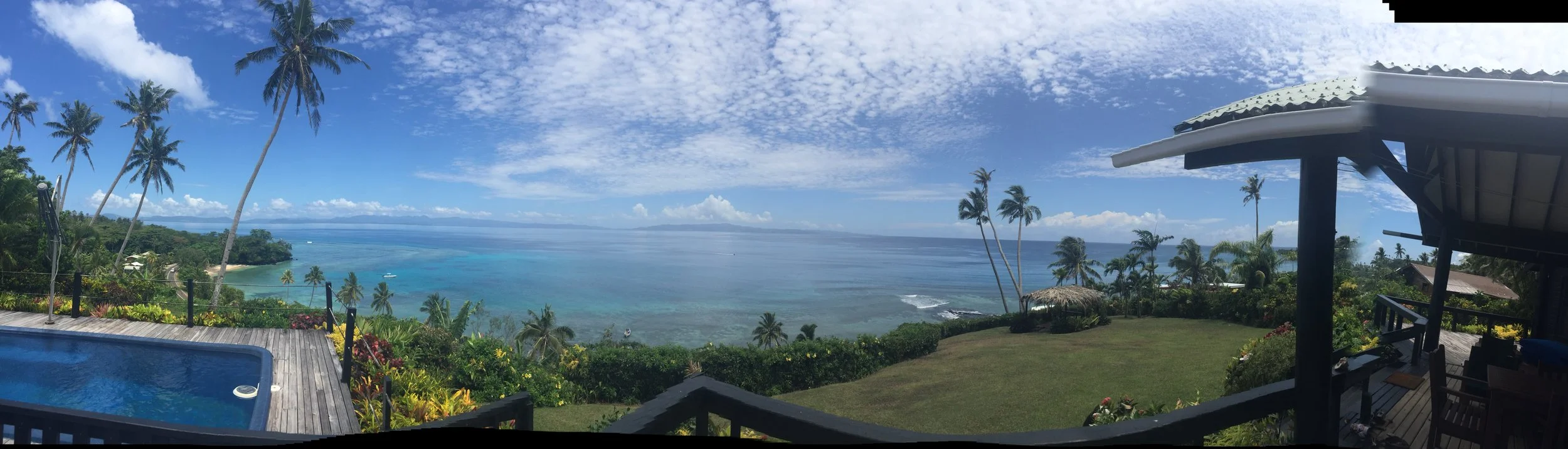 Ucuilagi, Taveuni Island Fiji - Panoramic view of the ocean with clear blue water, palm trees, and a partly cloudy sky. There is a pool, a grassy area, and the main house on the right side of the image. 