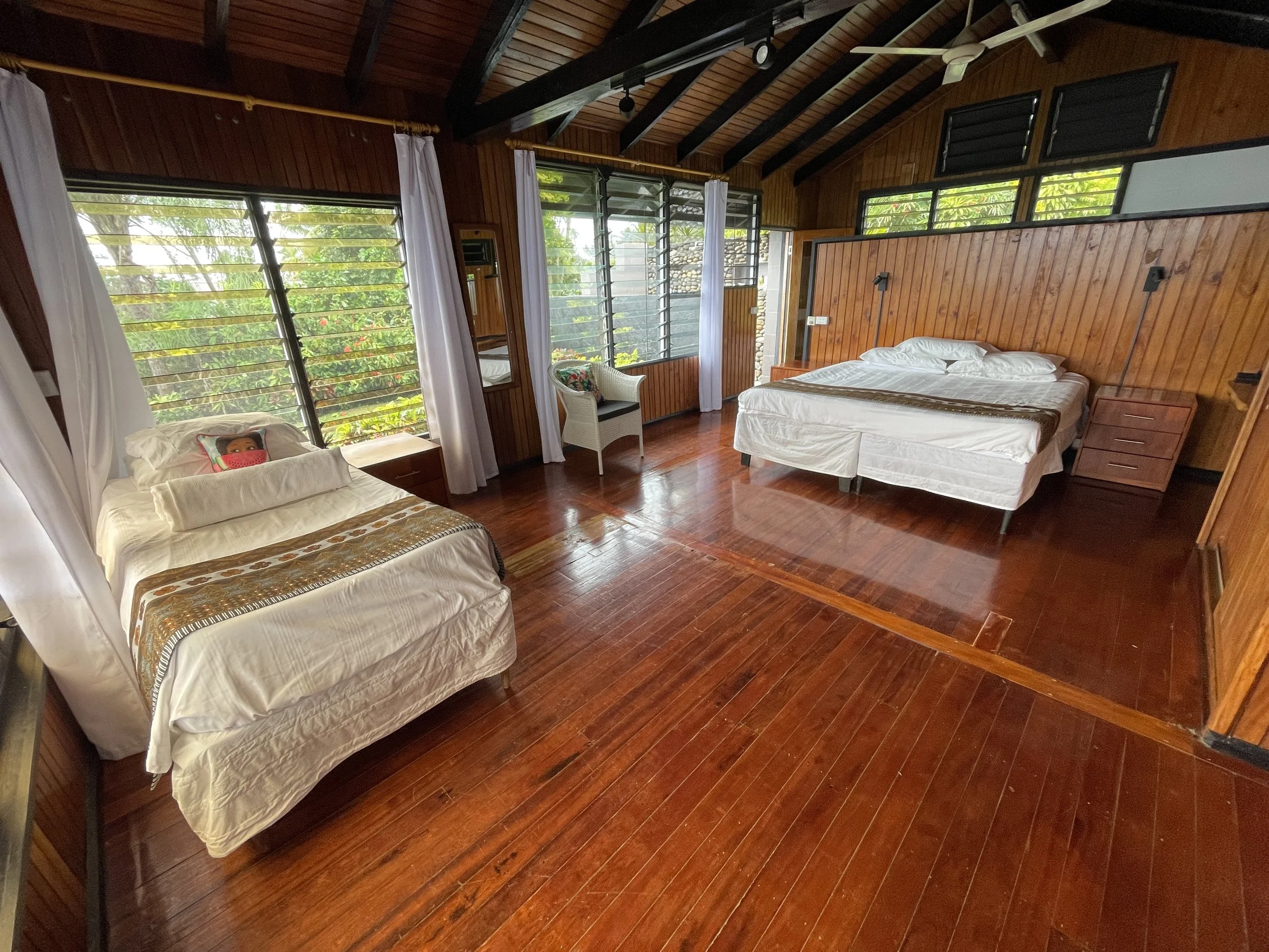 Ucuilagi, Taveuni Island Fiji - Master bedroom with two beds - king and single, a window with white curtains, and wooden walls and ceiling. Outside the window, lush green foliage is visible.