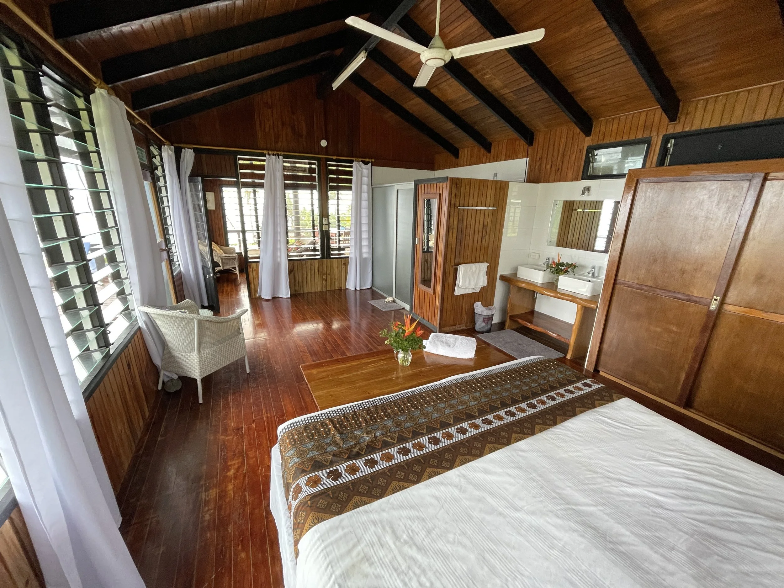 A cozy room with wooden walls and ceiling, featuring large windows with white curtains, a ceiling fan, a bed with a patterned bedspread, a small wooden vanity with a mirror and flower vase, and a wicker chair near the window.