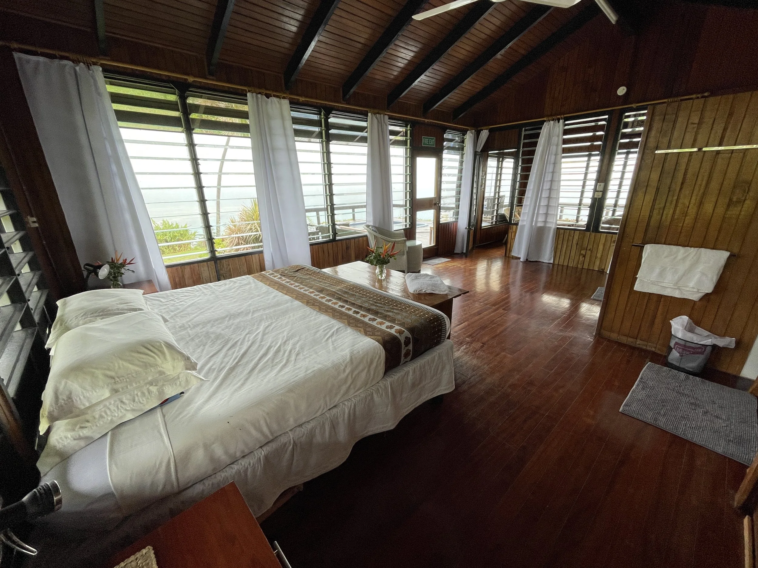 A cozy wooden interior with large windows, white curtains, a bed, a small table with a flower vase, and a chair.