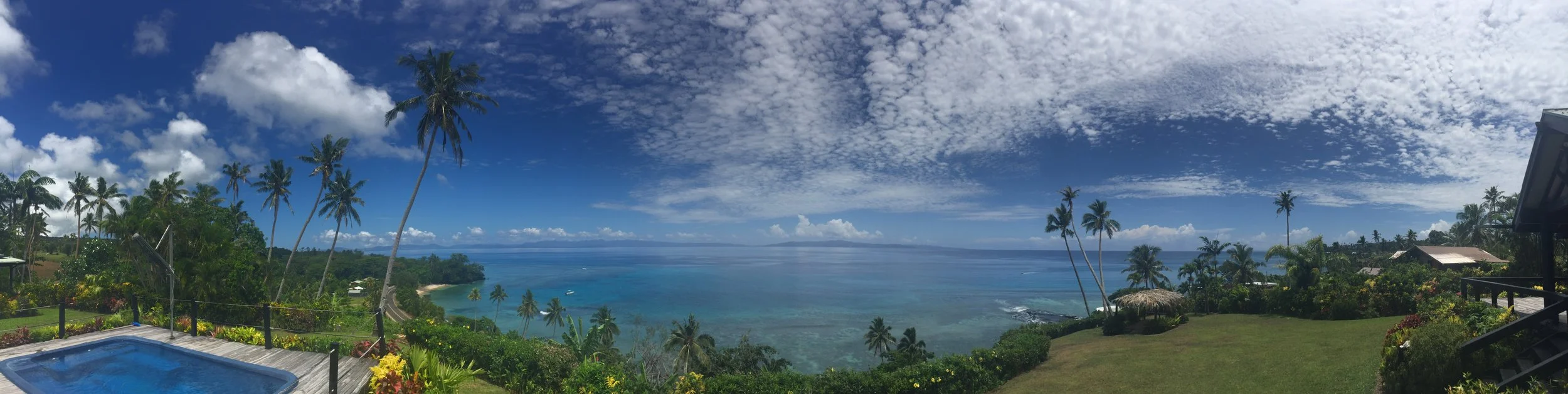 Ucuilagi, Taveuni Island Fiji - Panoramic view of a tropical beach and ocean from a hilltop with palm trees, a swimming pool, and green lawn.