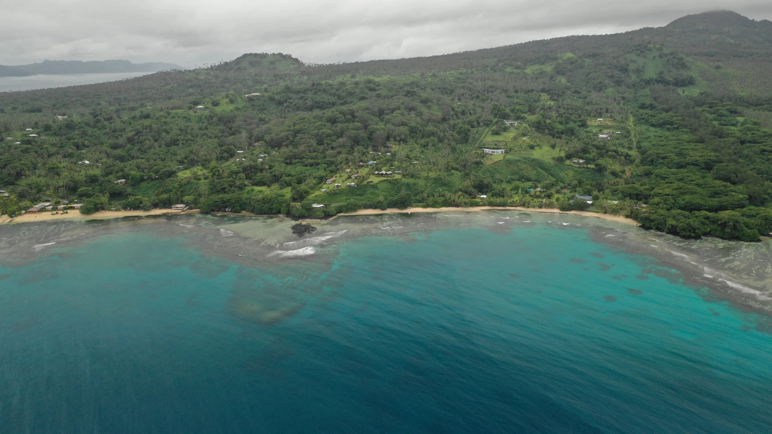 A tropical coastal landscape with green hills and a beach along turquoise ocean waters under a cloudy sky.