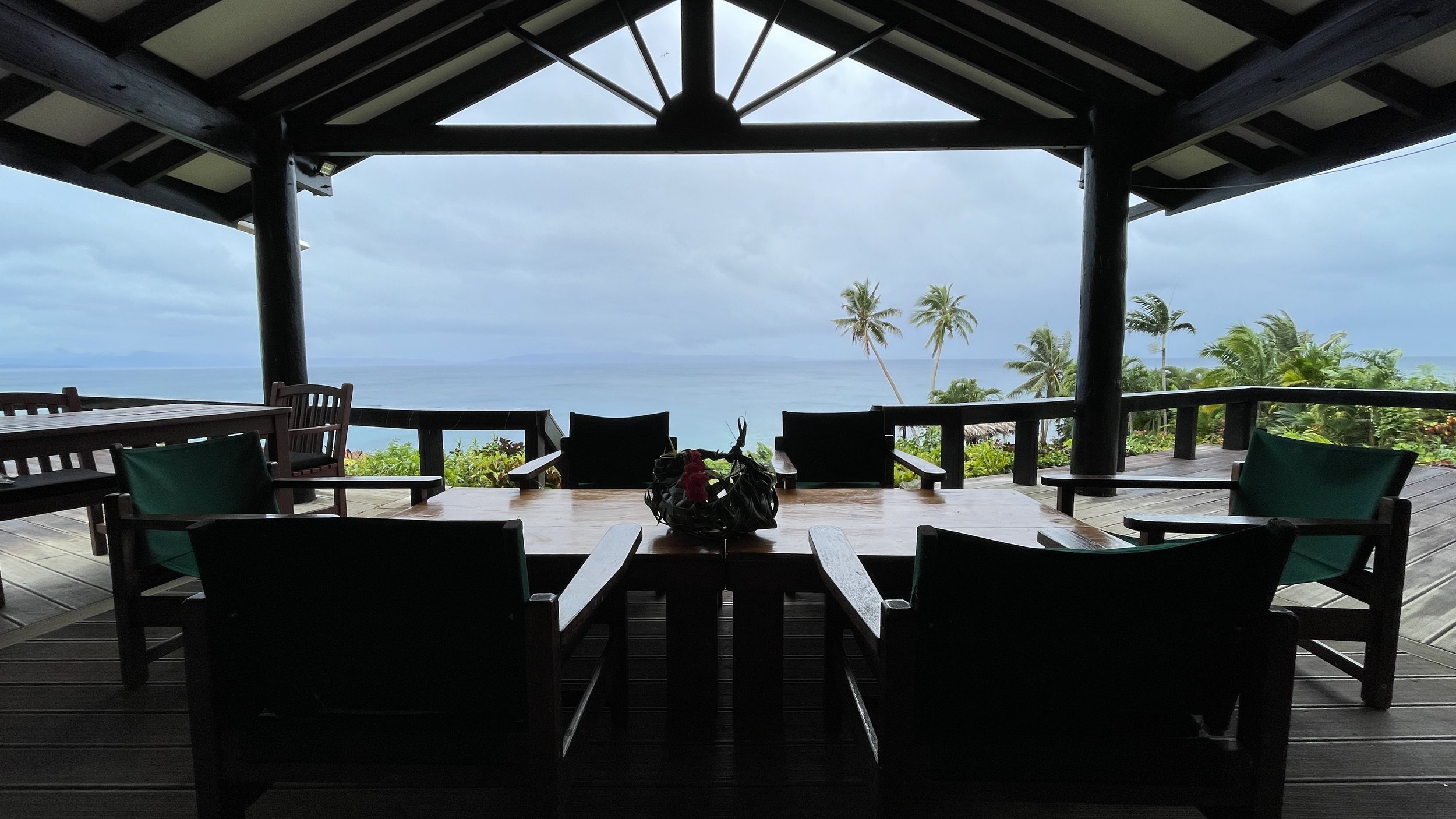 Overlooked wooden patio with chairs and table, overlooking a tropical landscape with palm trees and the ocean under a cloudy sky.