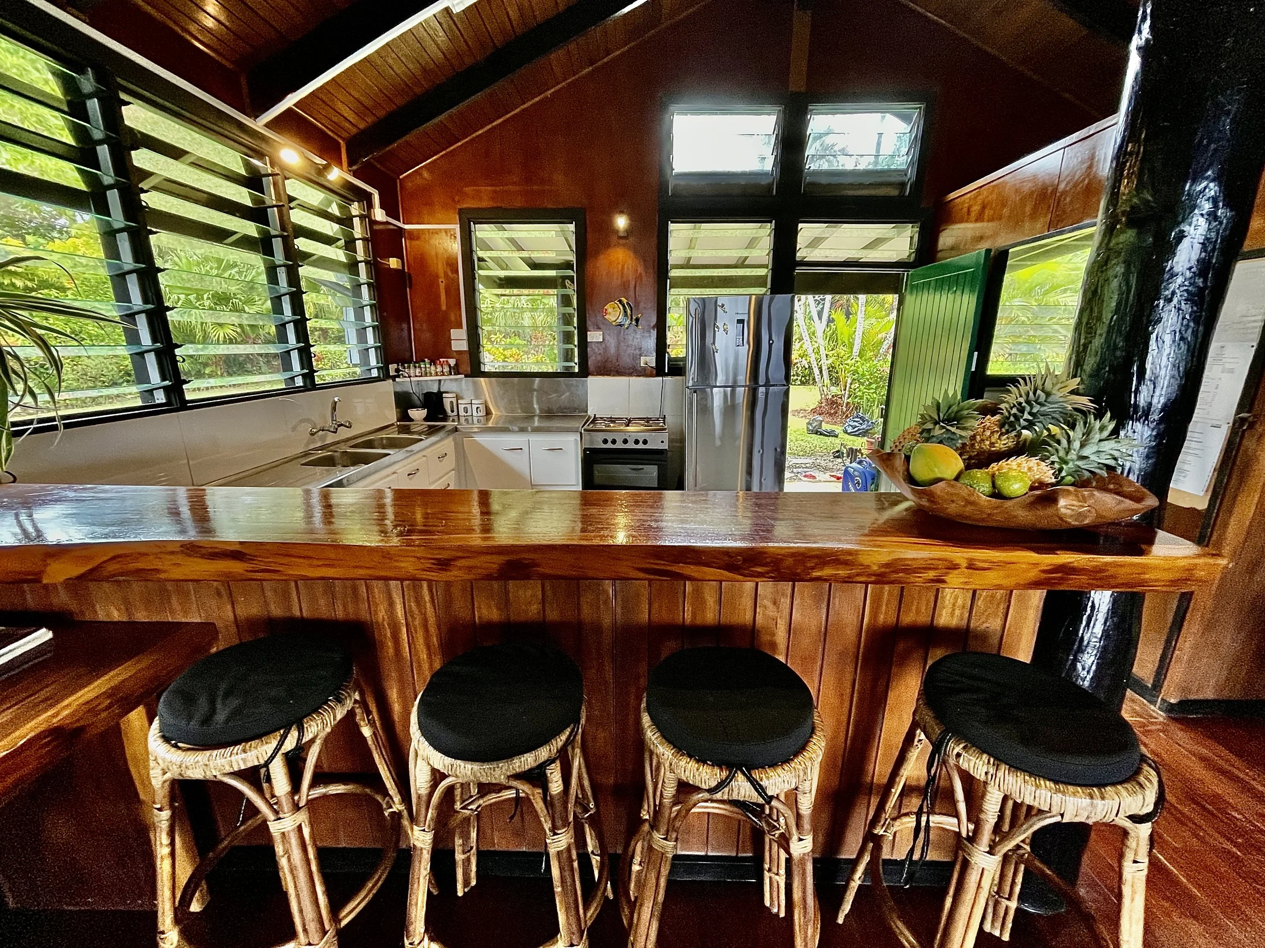A kitchen with a wooden countertop bar, four wicker bar stools with black cushions, a stainless steel refrigerator, and multiple windows bringing in greenery from the outside.