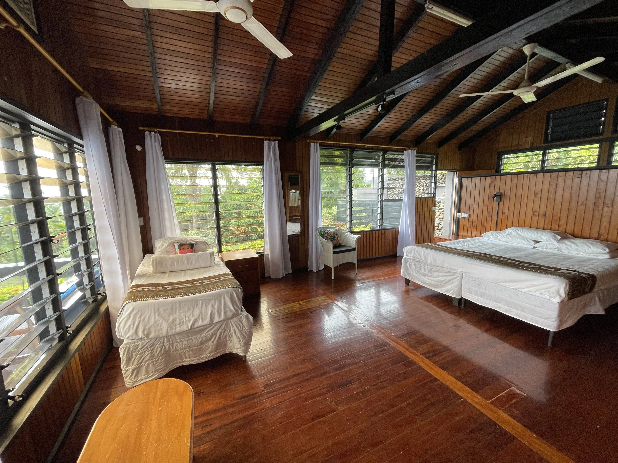 A bedroom with wooden walls and ceiling, featuring ceiling fans, large windows with white curtains, and beds with white linens.