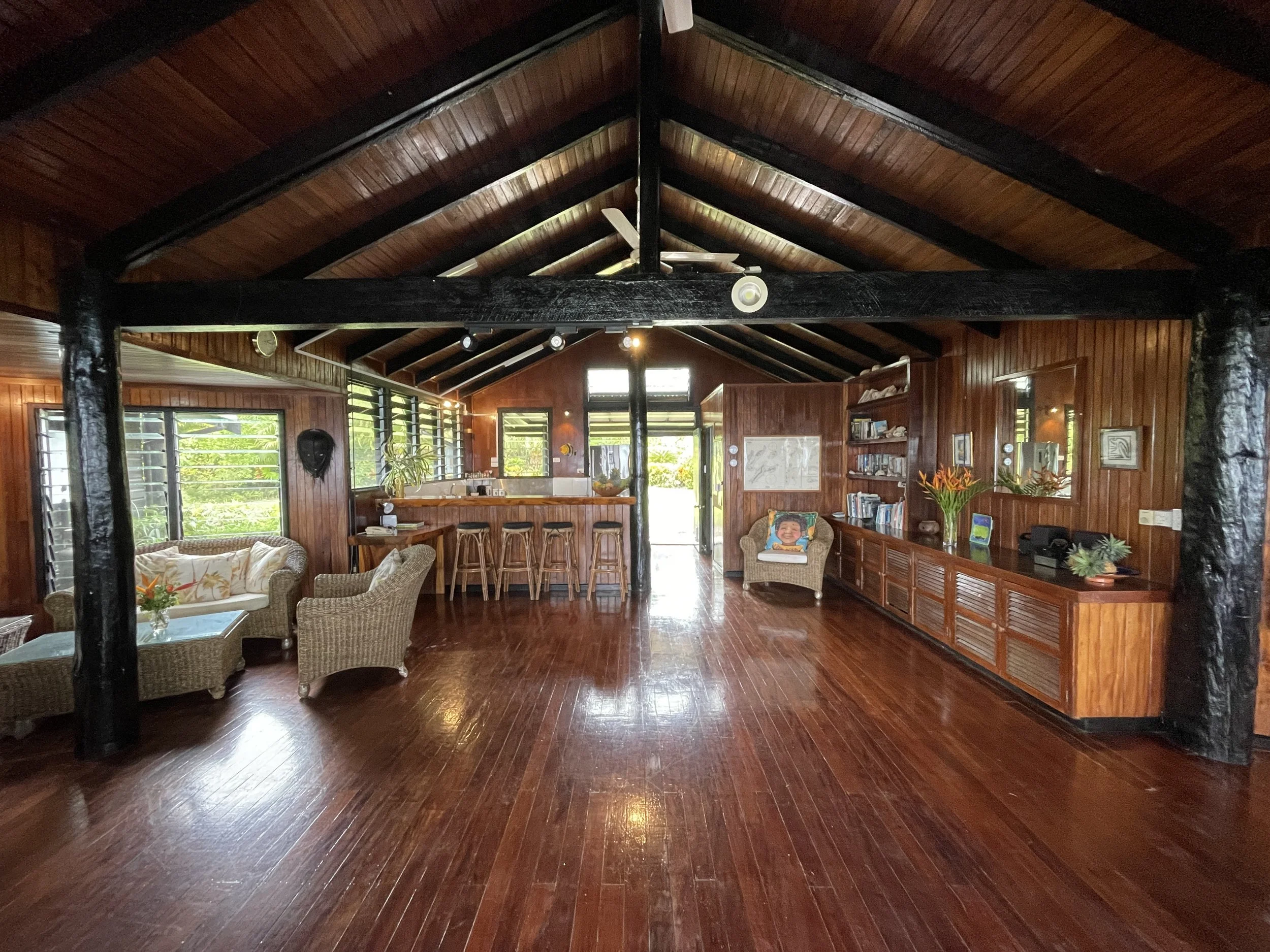 Spacious living room with wooden walls and ceiling, large windows with blinds, wicker furniture, bookshelf, and decorative plants.