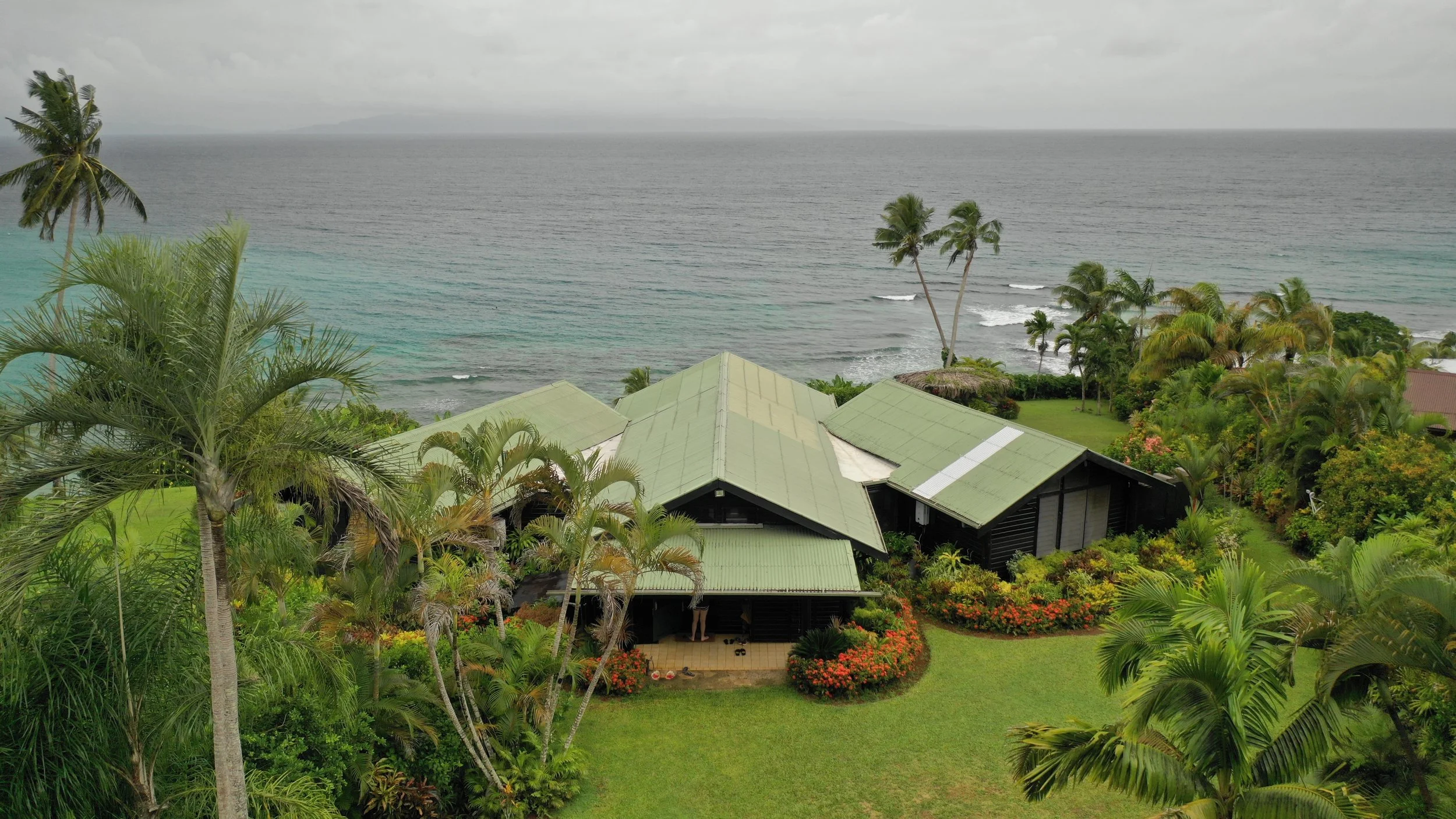 Ucuilagi, Taveuni Island Fiji - View from behind the main tropical clifftop house overlooking Somosomo Strait surrounded by lush greenery and palm trees, with the ocean and cloudy sky in the background.