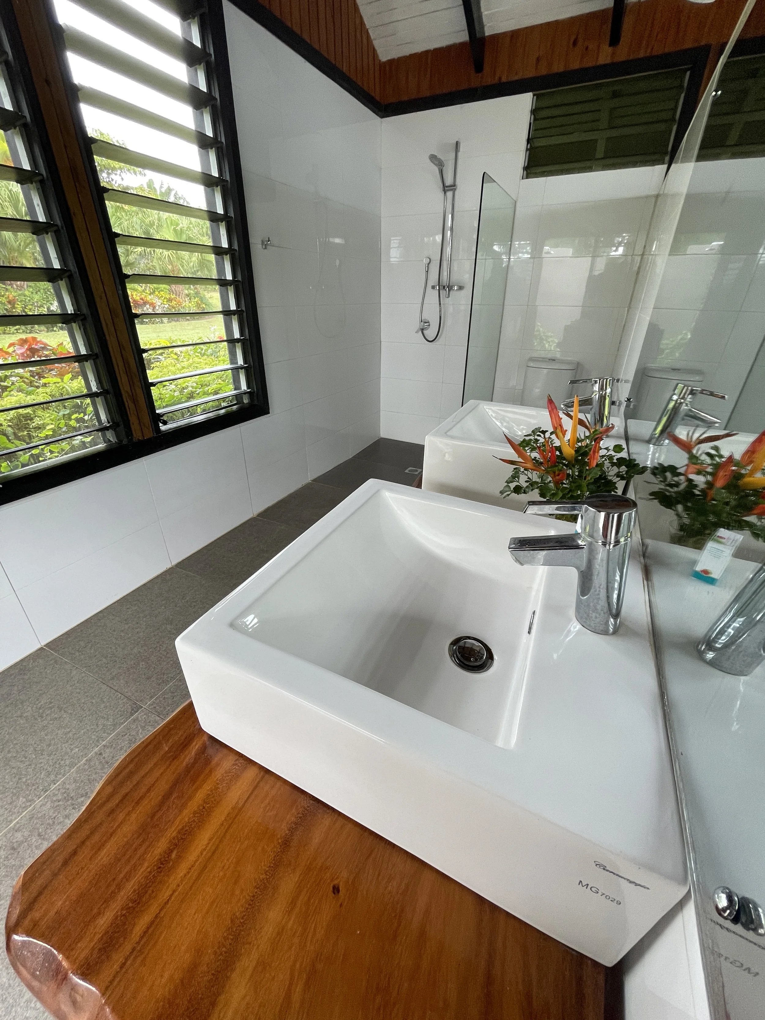 View of a modern bathroom with a white sink, faucet, a mirror, a shower area with a showerhead, a window with louvers, and a vanity with a bouquet of flowers.