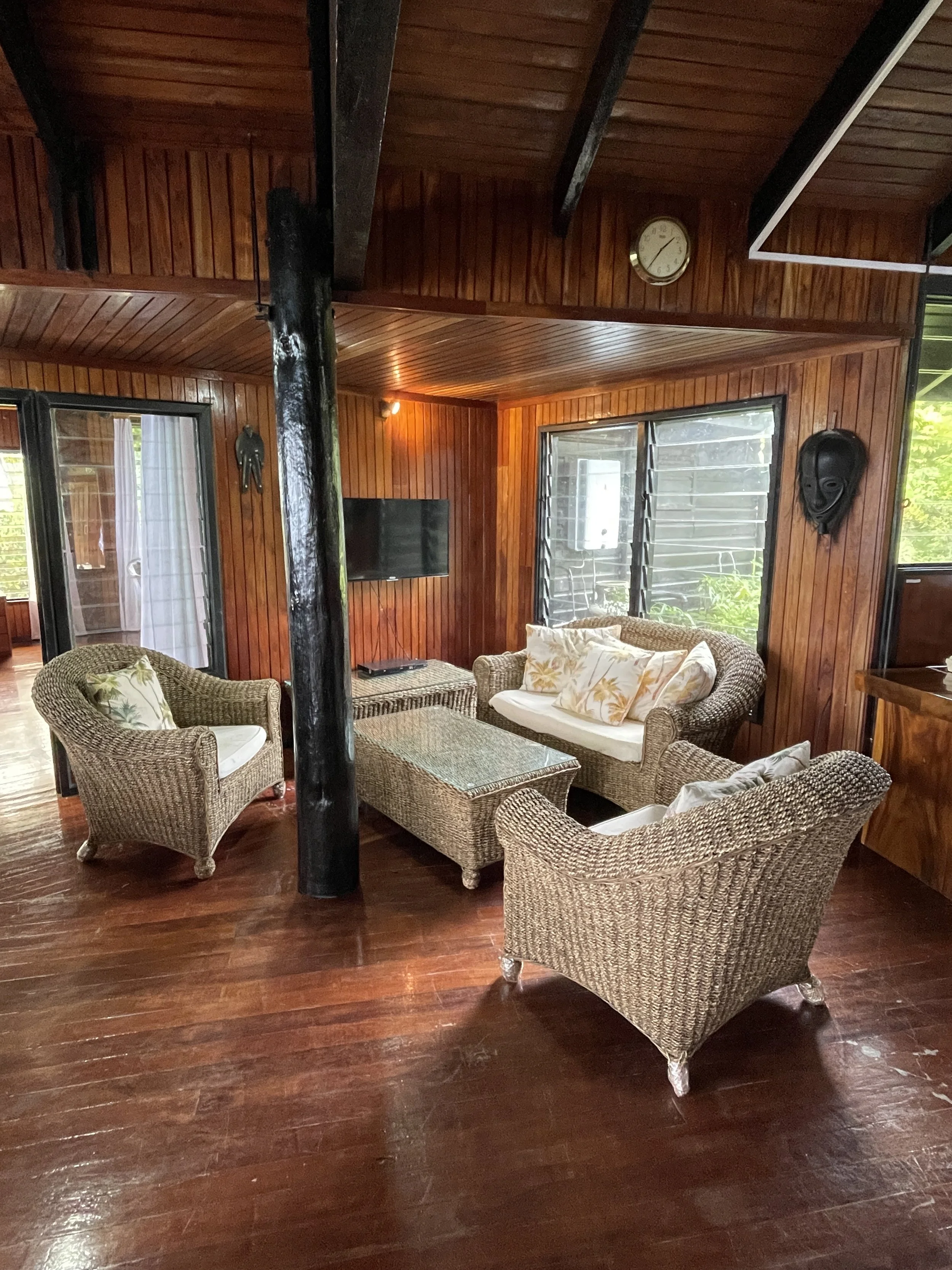 A cozy indoor living area with wooden walls and ceiling. There are wicker chairs with cushions, a glass-top coffee table, and a wall-mounted TV. Windows with shutters and a clock on the wall are visible.
