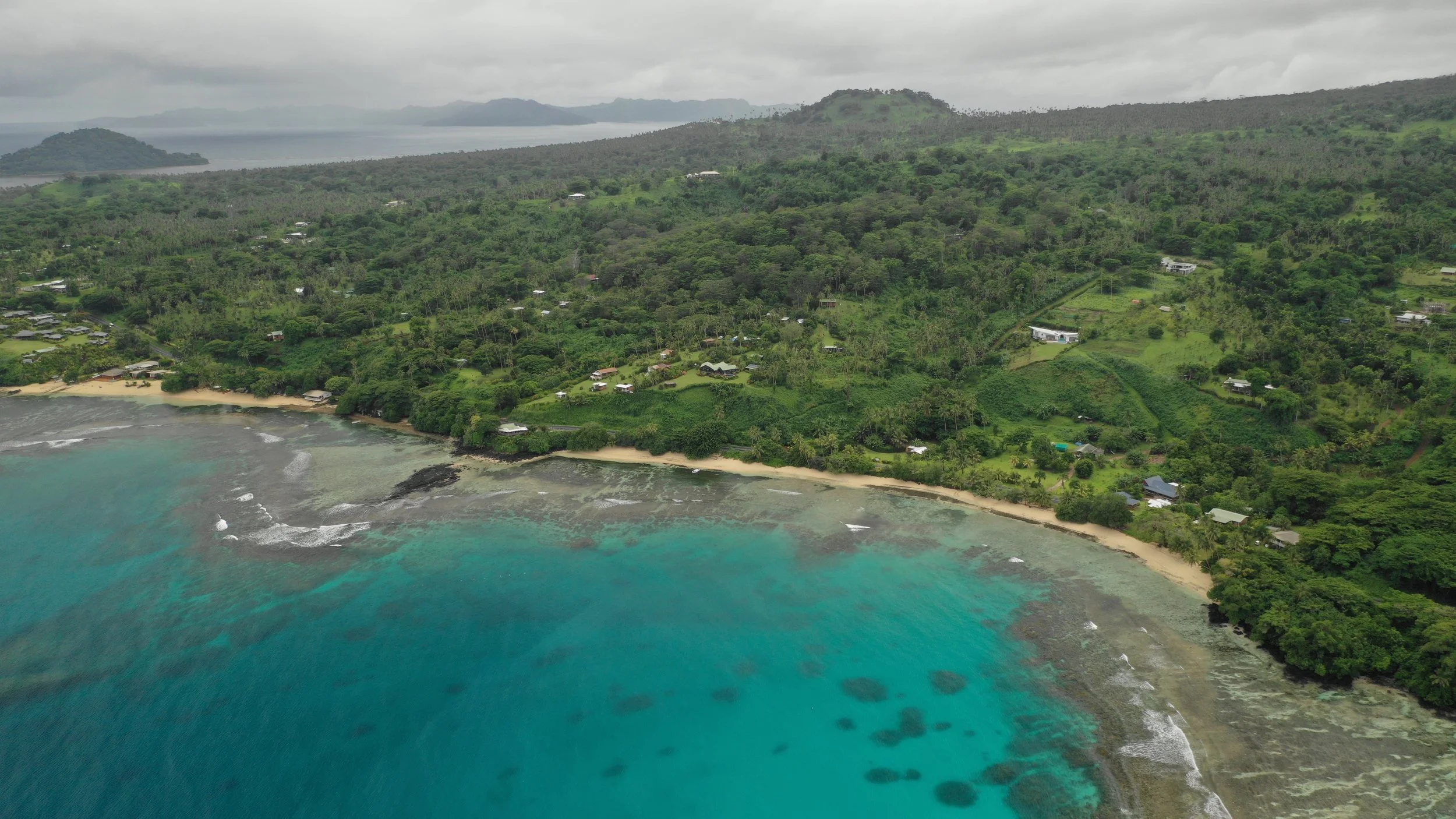 An aerial view of a lush green coastline with a sandy beach, turquoise water, and scattered houses nestled among trees, with hills and mountains in the background under cloudy skies.