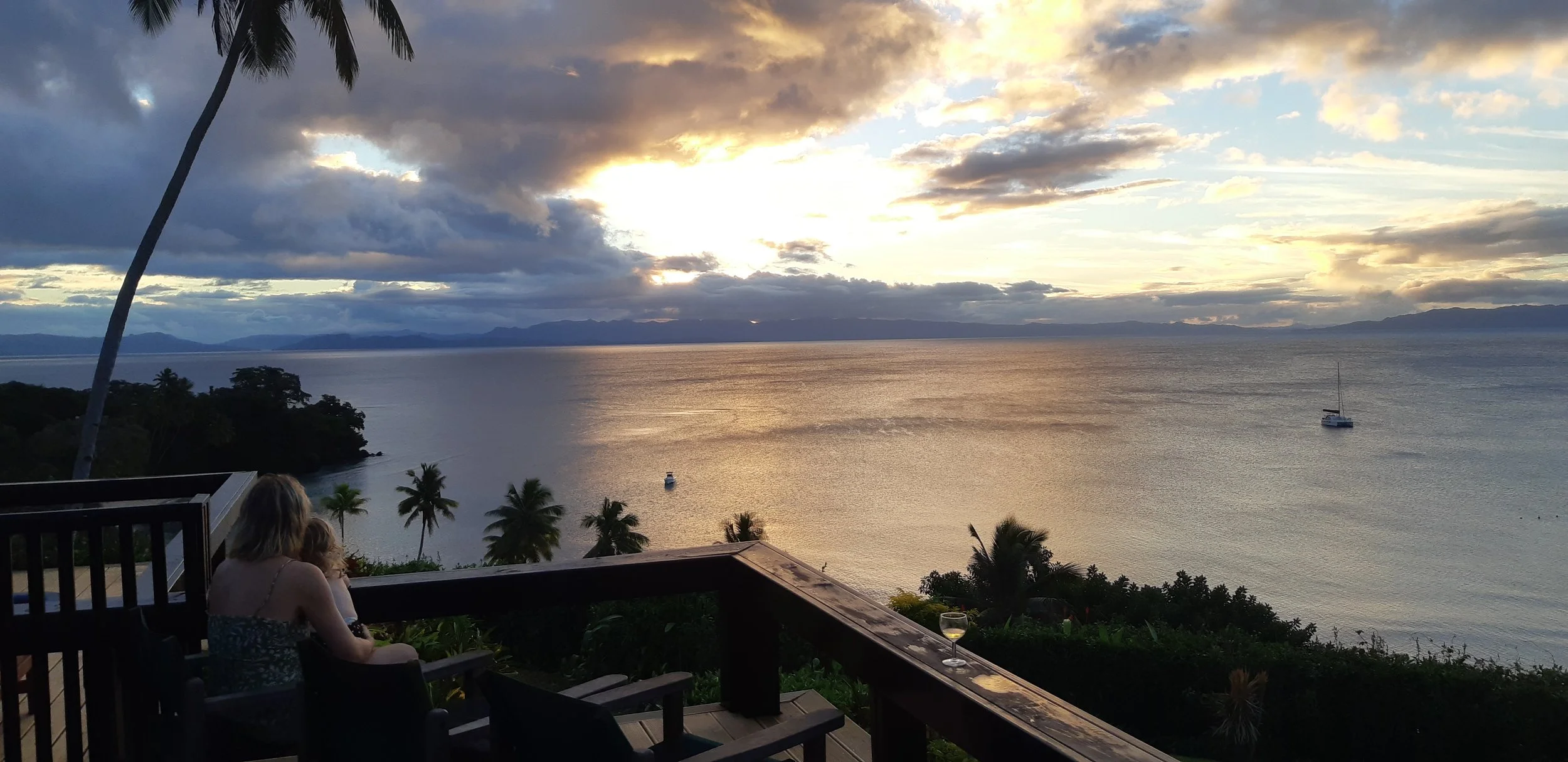 A sunset over the ocean with dark clouds and a golden sky. In the foreground, there are palm trees, a woman with a child sitting on a balcony, and a glass of wine on the railing. Two boats are visible on the water.