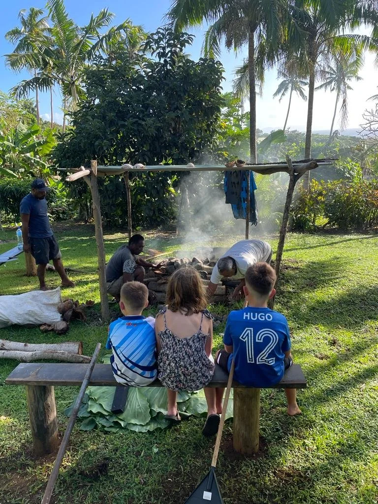 Ucuilagi, Taveuni Island Fiji - Three children sitting on a wooden bench watching a group of adults cooking Fijian meal Lovo over fire. The setting is outdoors among lush green trees and grass.