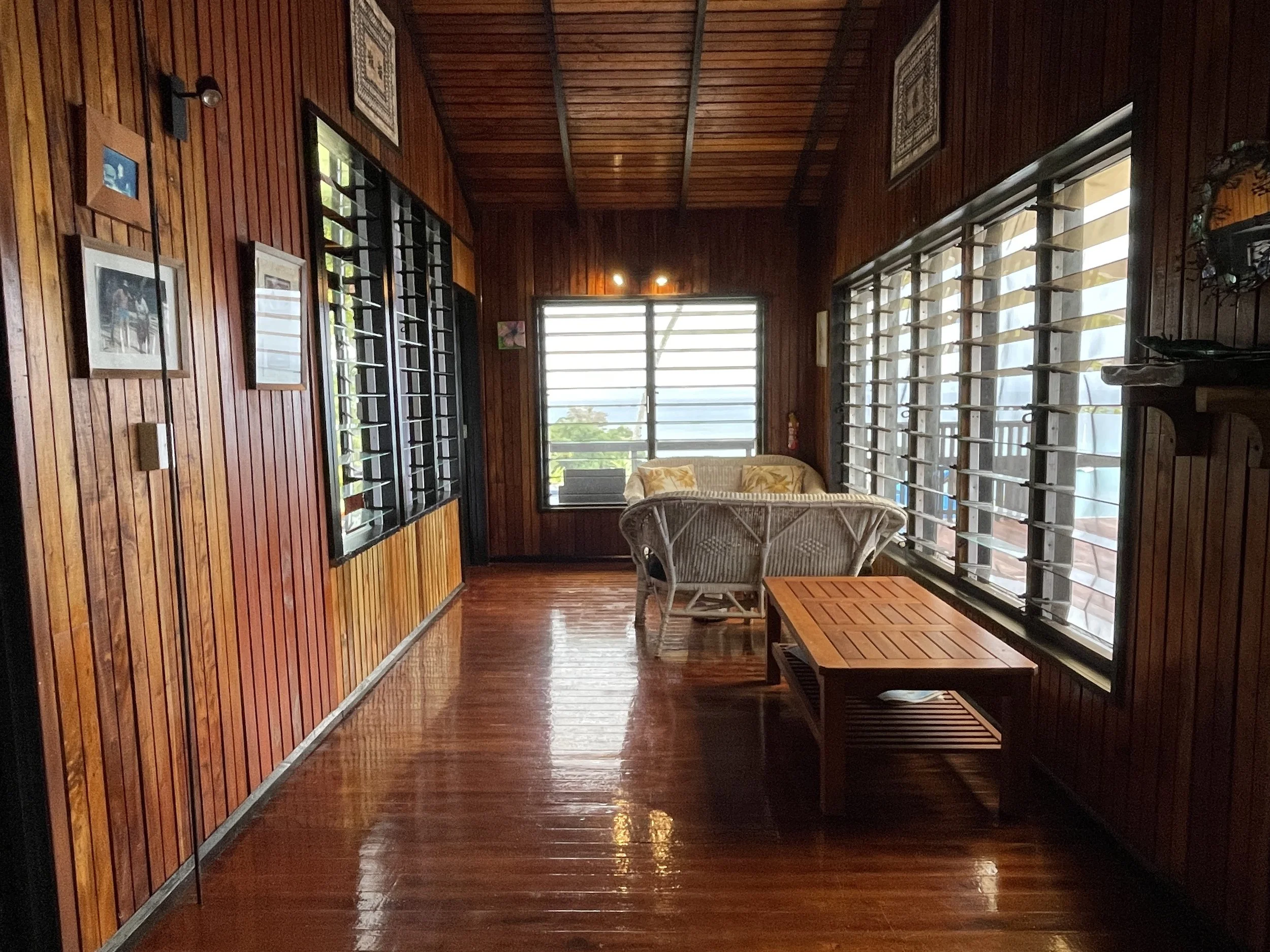 A cozy wooden porch or sunroom with large windows, wicker chairs, a small table, and framed photographs on the walls.
