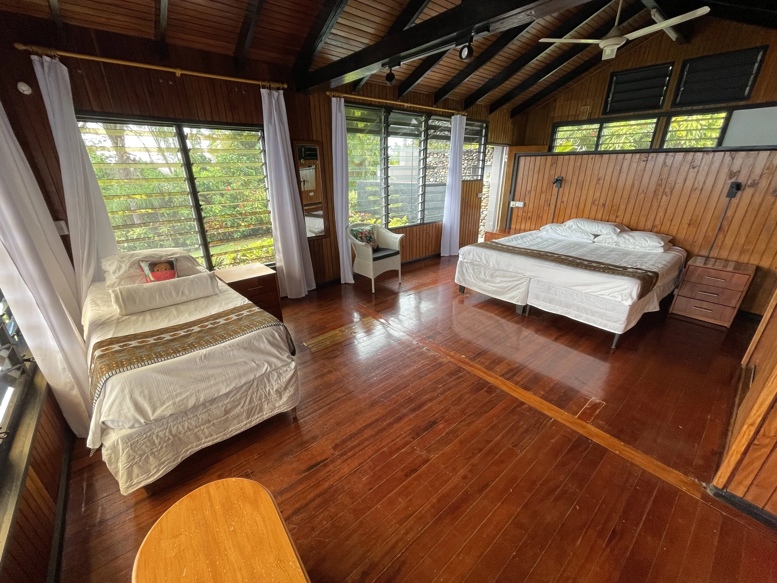 A bedroom with wooden walls and ceiling, two beds with white linens, a small wooden side table, a wicker chair with a colorful cushion, large windows with horizontal louvers and white curtains, and a polished wooden floor.