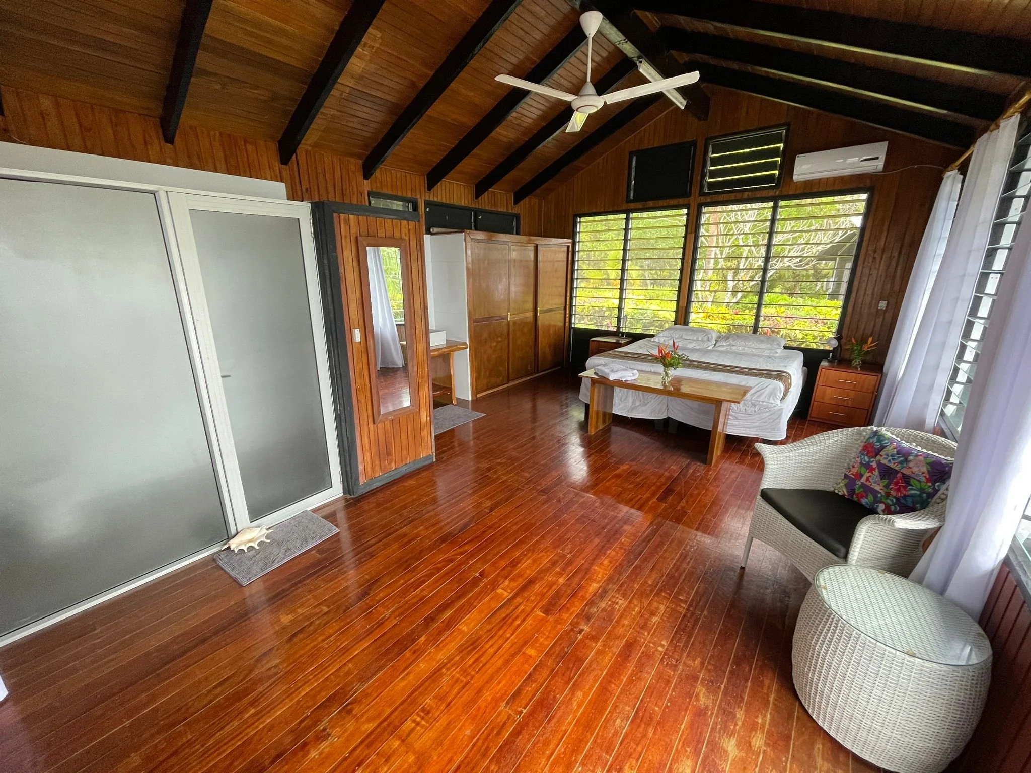 Interior of a bedroom with wooden walls and ceiling, a white bed with pillows and a floral pillow, a small wooden table with flowers, a wicker chair, windows with greenery outside, a ceiling fan, air conditioner, and sliding door.
