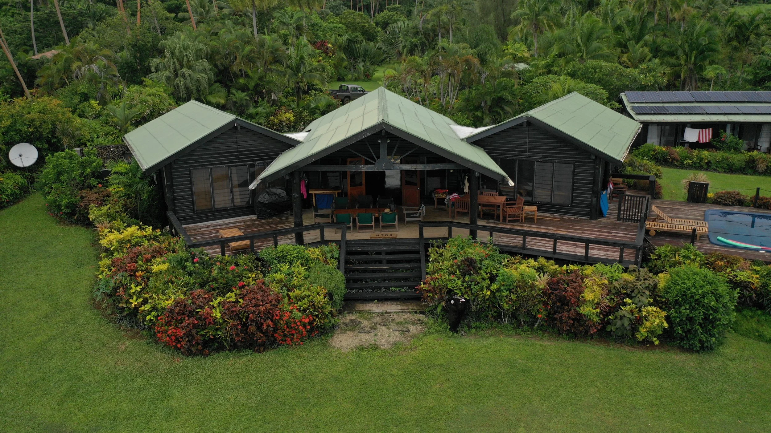 Aerial view of a black house with a gray roof, surrounded by lush green tropical plants and trees, with a wooden deck and a small swimming pool.