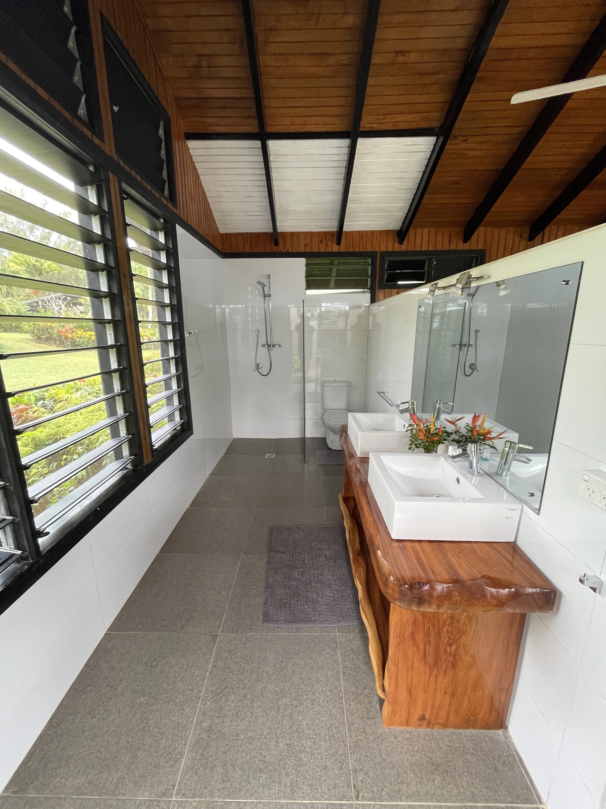 A bathroom with white tiled walls, a wooden countertop with two white sinks, a large mirror, and a gray floor mat. There are two windows with black frames and louvered glass panes, one open and one closed. The ceiling is wooden with black beams and a