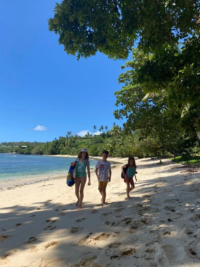 Vacala Bay, Ucuilagi, Taveuni Island Fiji - Three children walking along a sandy tropical beach near the water's edge, with trees and greenery overhead and a clear blue sky in the background.