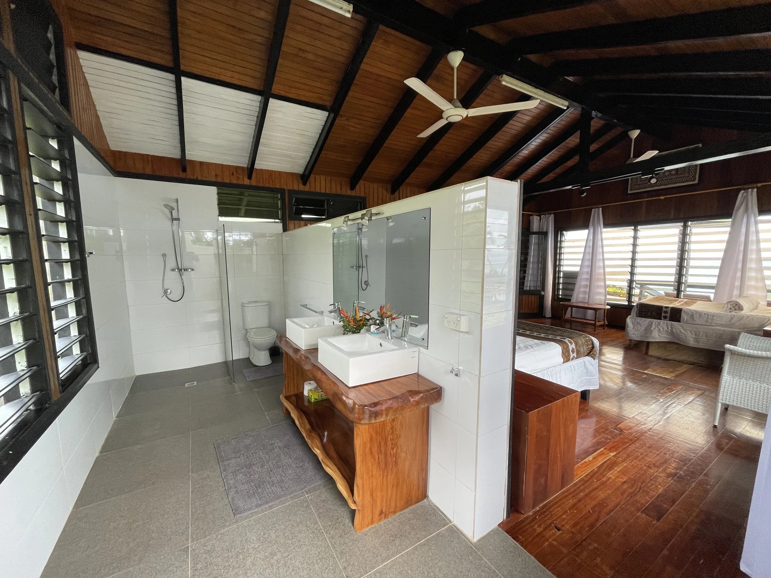 A room with a bathroom and a bedroom separated by a partial wall. The bathroom has white tiles, a toilet, a sink with a mirror, and a shower. The bedroom features wooden floors, a bed with a patterned comforter, and windows with curtains. The room ha