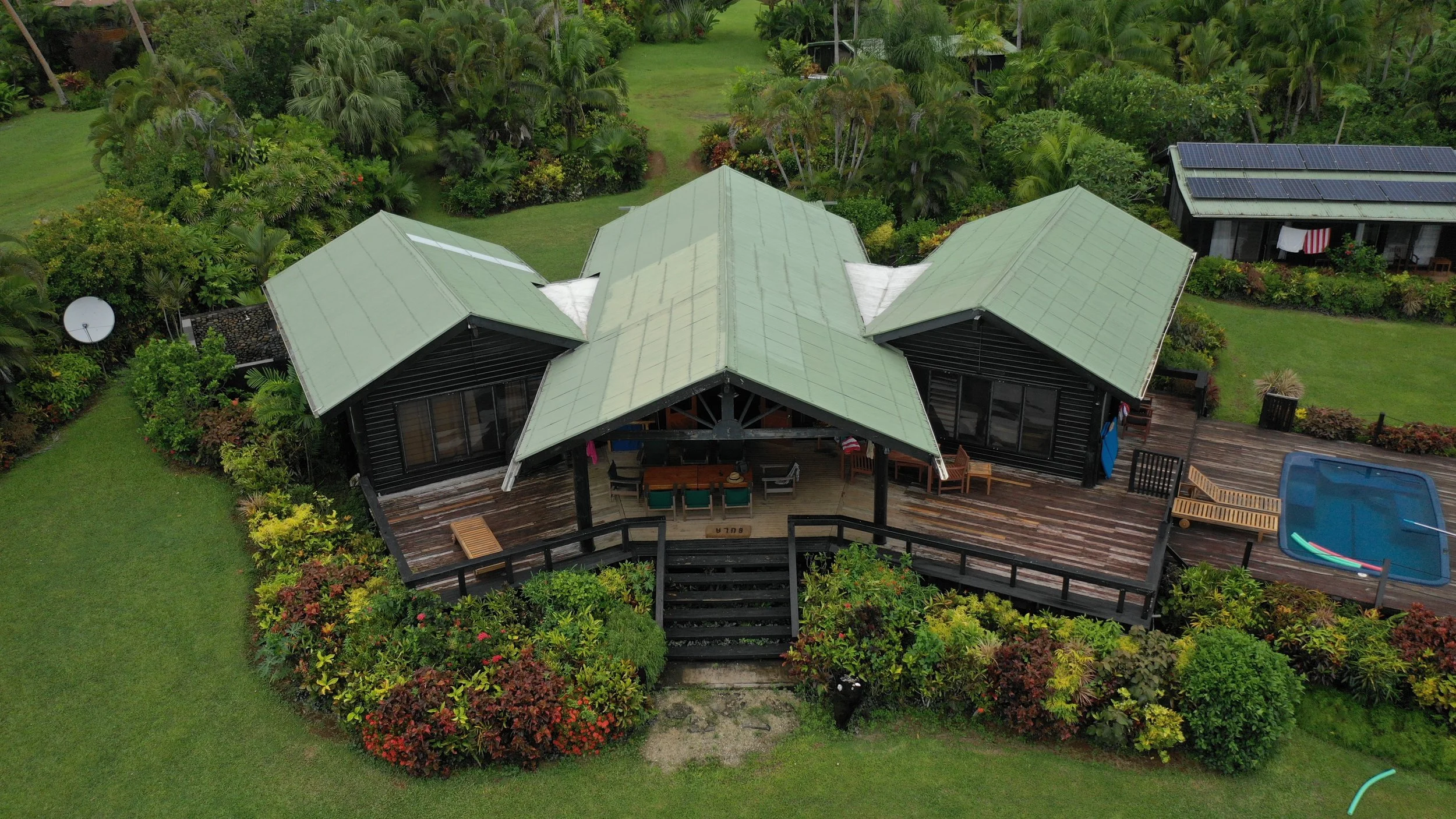 Ucuilagi, Taveuni Island Fiji - An aerial view of a house with a green metal roof, surrounded by lush tropical vegetation, a wooden deck with outdoor furniture, a small pool, and a satellite dish on the left side.
