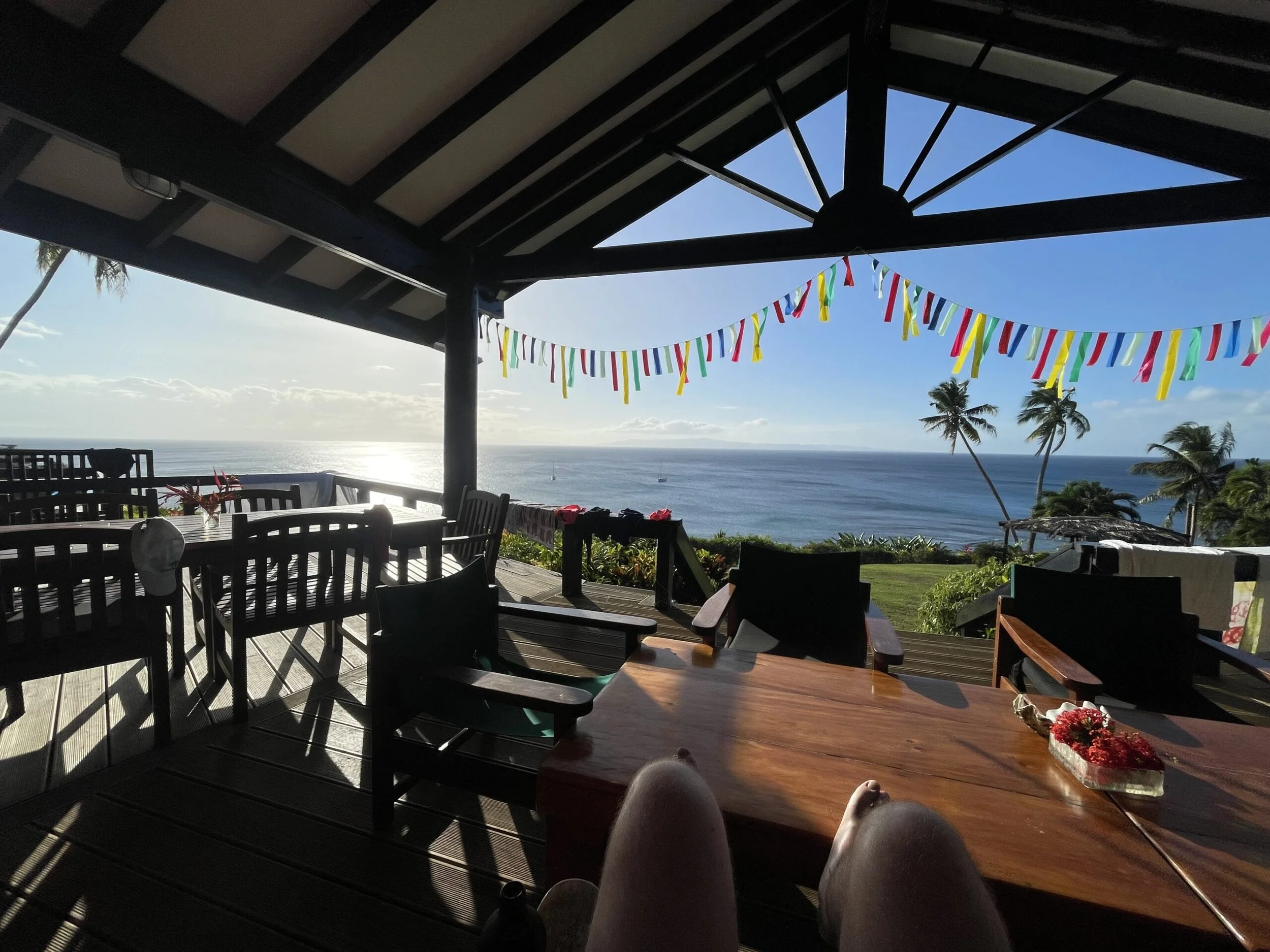 Ucuilagi, Taveuni Island Fiji - A covered outdoor patio overlooking the ocean with palm trees in the distance. There are tables and chairs, and colorful streamers hanging from the roof.