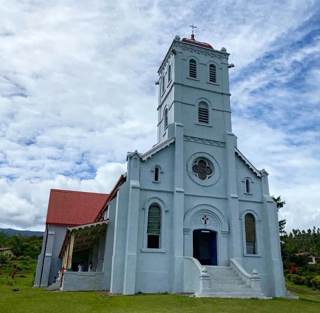 Light blue church with red roof, arched windows, and steeple under a partly cloudy sky, surrounded by green grass and distant trees.