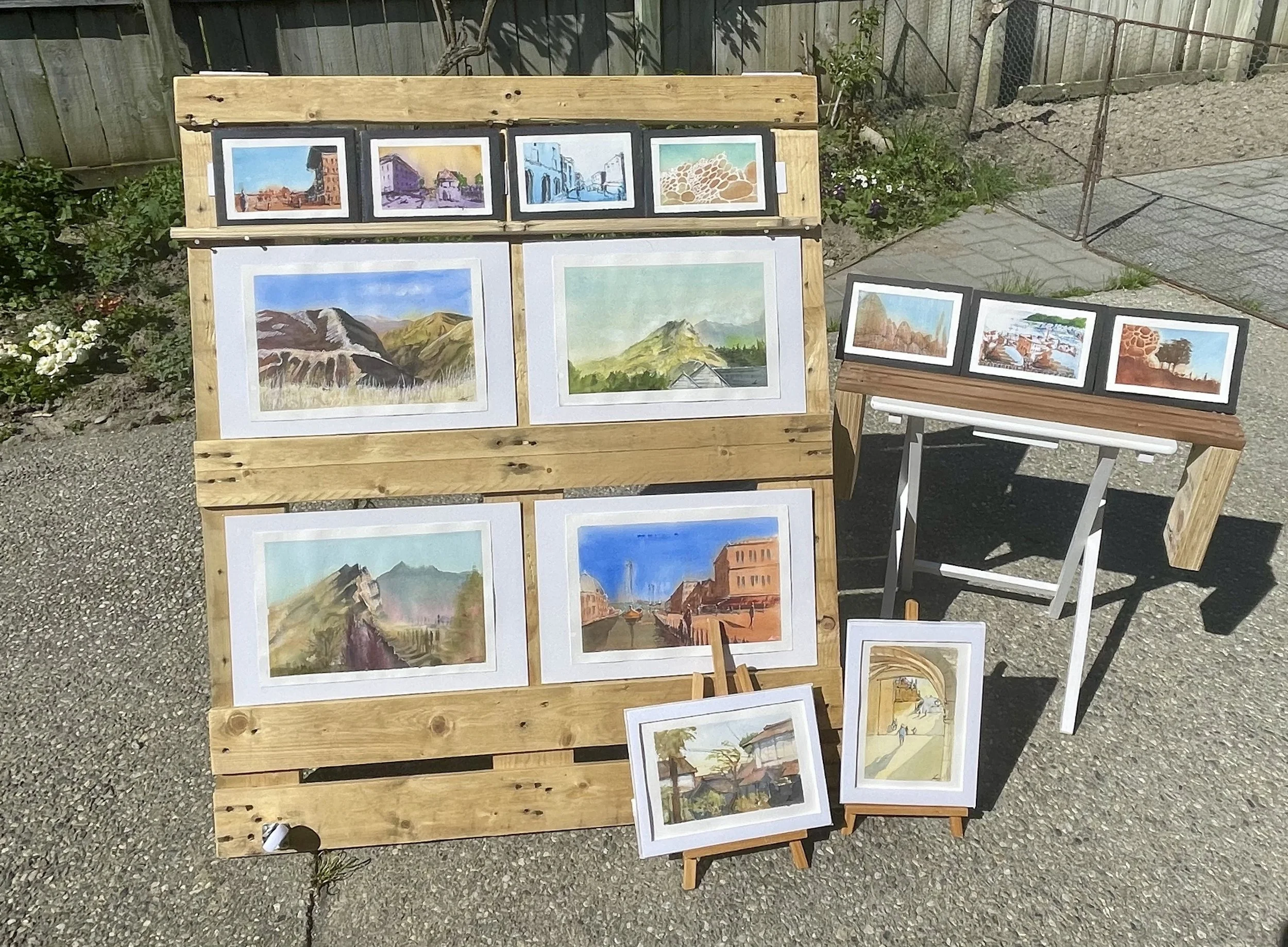 Outdoor art display with watercolor paintings of landscapes and buildings on a wooden and white stand, and additional artwork on small easels nearby, with a garden and fence in the background.