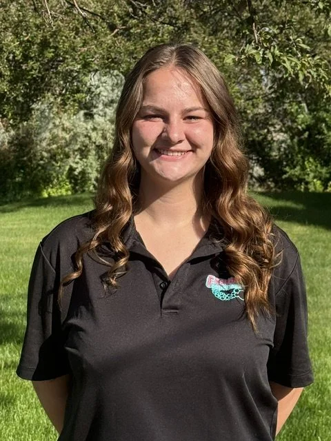 Young woman with long wavy brown hair smiling outdoors in front of green trees and grass, wearing a black polo shirt with a logo on the left chest.