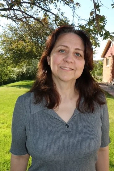 A woman standing outdoors on a sunny day in a grassy area with trees and a building in the background.