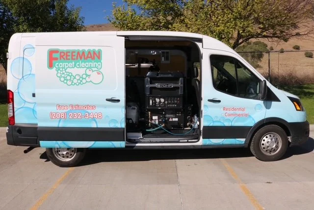 A white van with the logo 'Freeman Carpet Cleaning' and contact information on the side, parked outdoors with trees and a hill in the background.