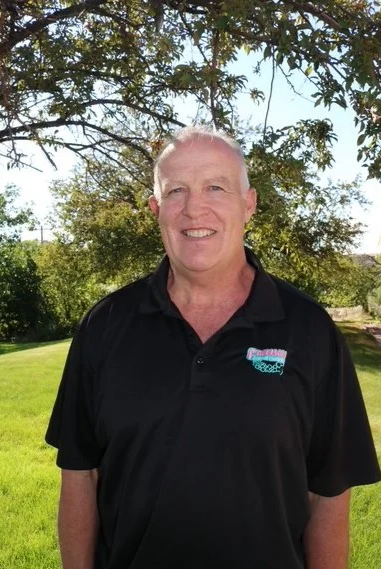 A man with short, light-colored hair smiling outdoors under a tree, wearing a black collared shirt with a small logo.