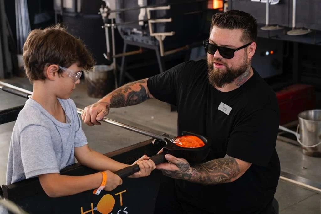 A man with tattoos and sunglasses is handing a bowl of orange-colored food to a young boy wearing glasses and a gray t-shirt, at an indoor event or food stall.