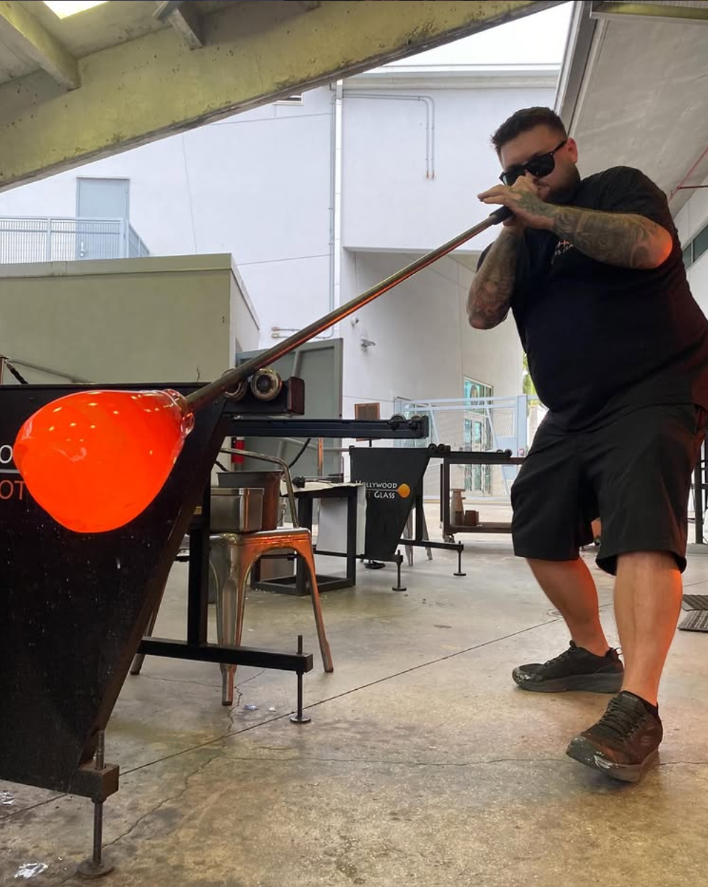 A glassblower inflates molten glass using a long blowpipe in a studio. The glass is glowing red-orange, indicating it's very hot. The artisan wears dark clothing and sunglasses, standing in front of a workbench with tools, and is in a focused pose.