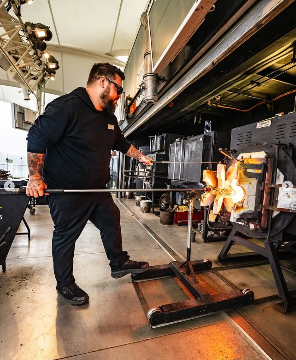 A man with a tattooed arm and beard, wearing black clothes and sunglasses, standing in front of a glassblowing furnace in a glass studio, using a long metal rod to shape molten glass.