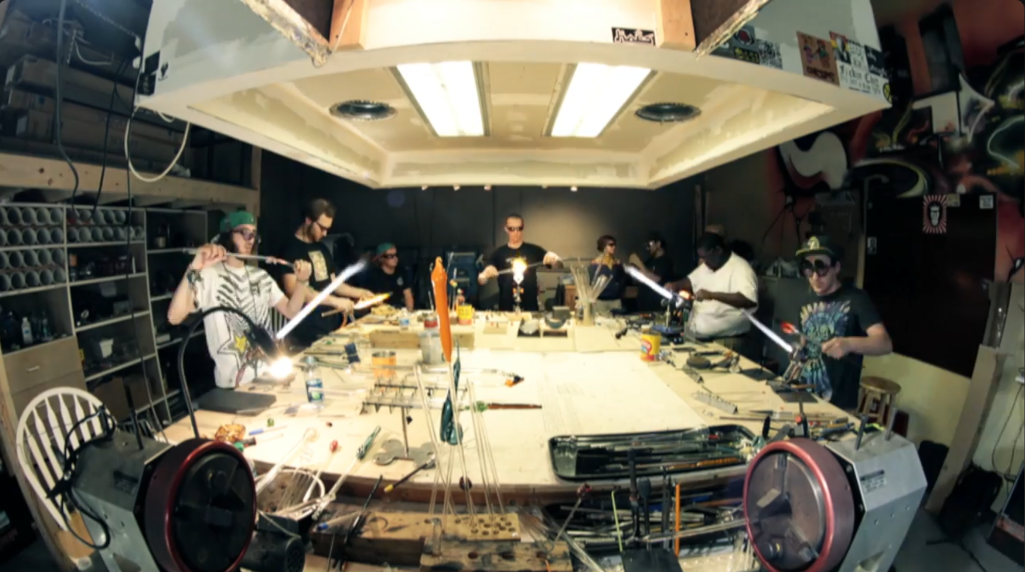 A group of people working at a large table in a workshop, with various tools and equipment, some using torches, and a ceiling with recessed lighting.